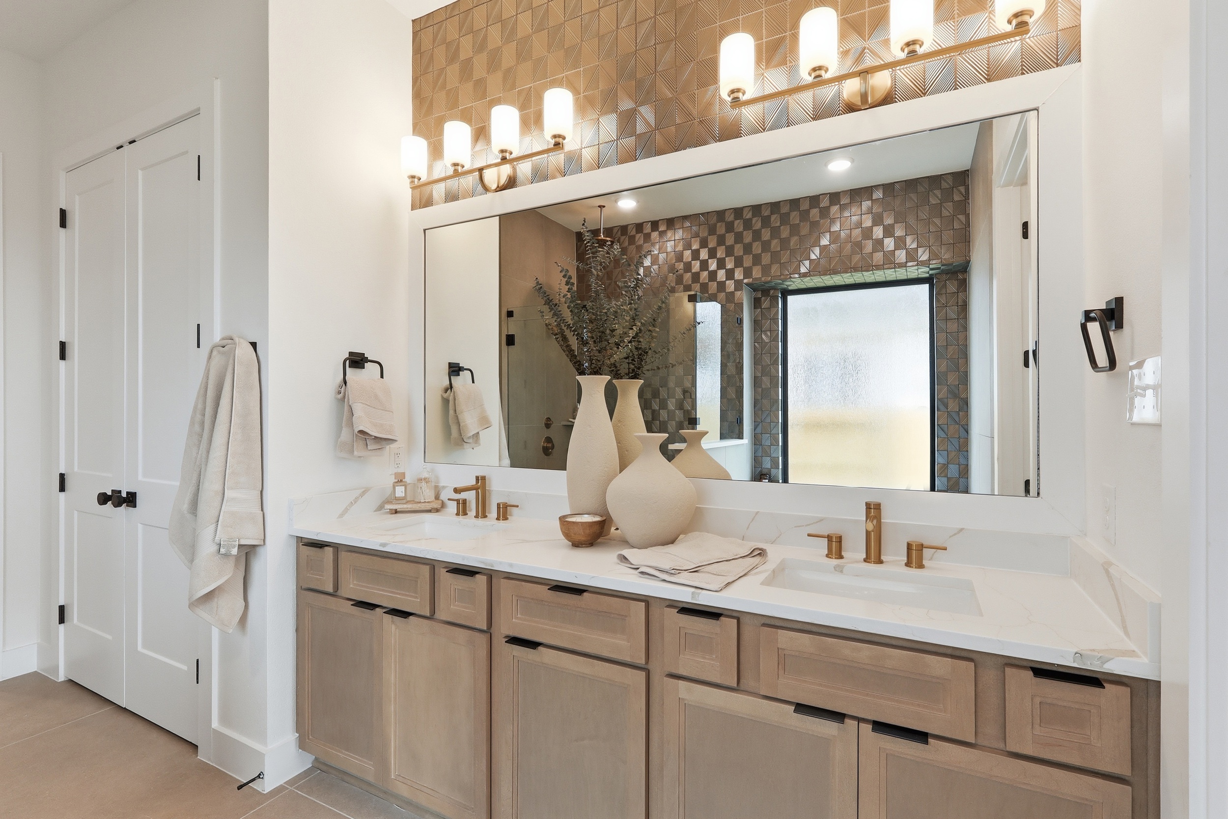 Modern bathroom with a double sink vanity, light wood cabinets, gold fixtures, large mirror, wall-mounted lights, neutral towels, and decorative vases. Patterned tile wall and frosted glass shower in the background.