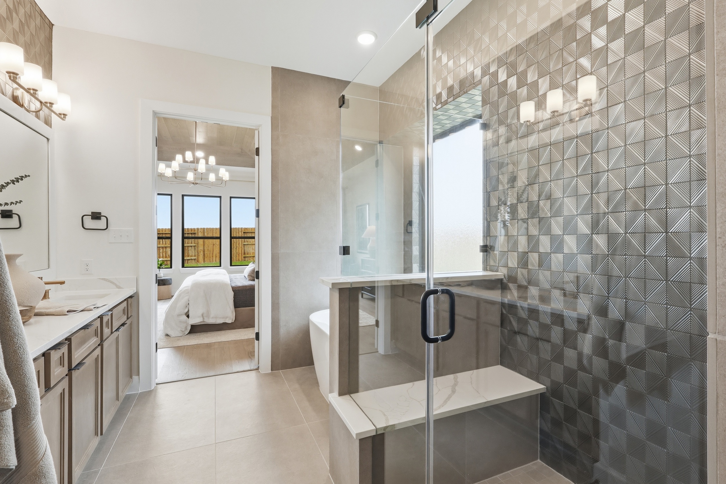 Modern bathroom with a glass-enclosed shower, geometric tiled accent wall, double sink vanity with towels, and view into a bedroom with large windows and a fenced backyard.