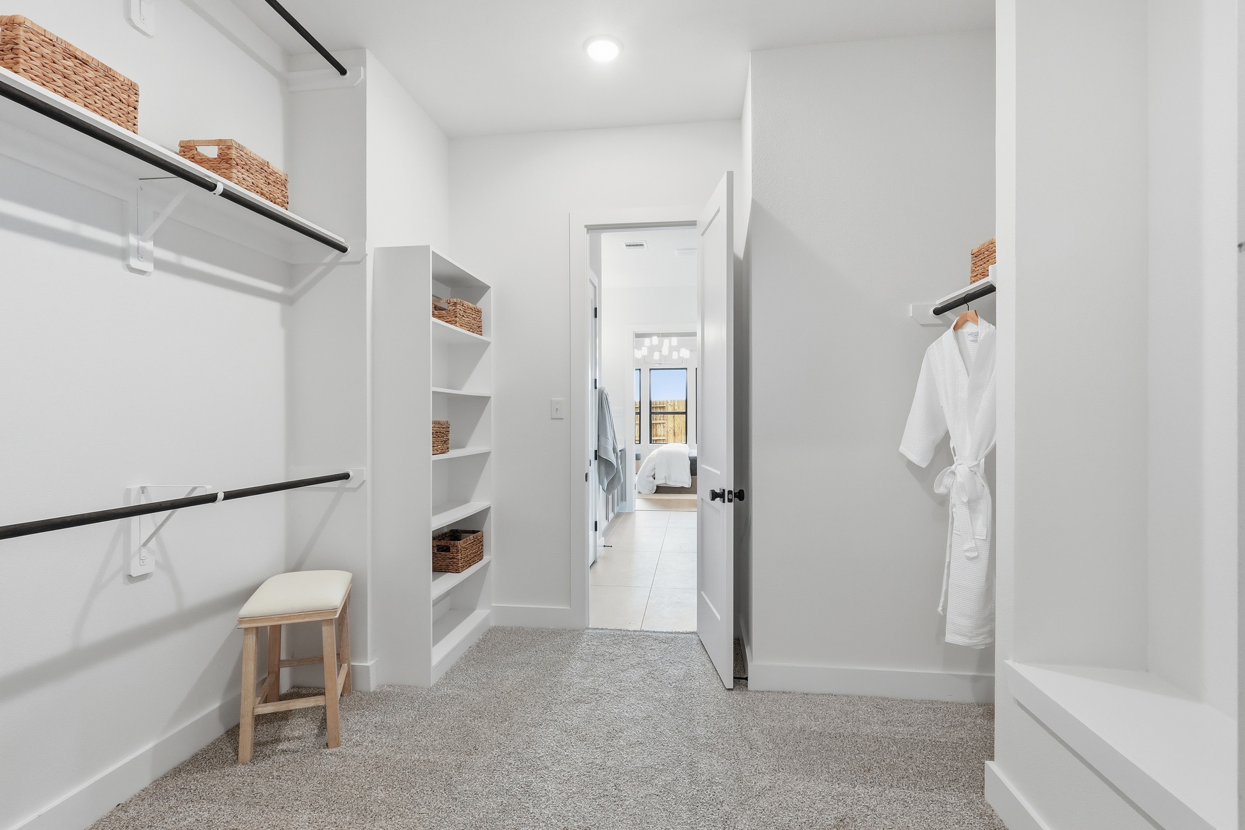 Spacious walk-in closet with shelves and hanging rods, wicker baskets for storage, a white robe, a small bench, and an open door leading to a bright bedroom with a large window.