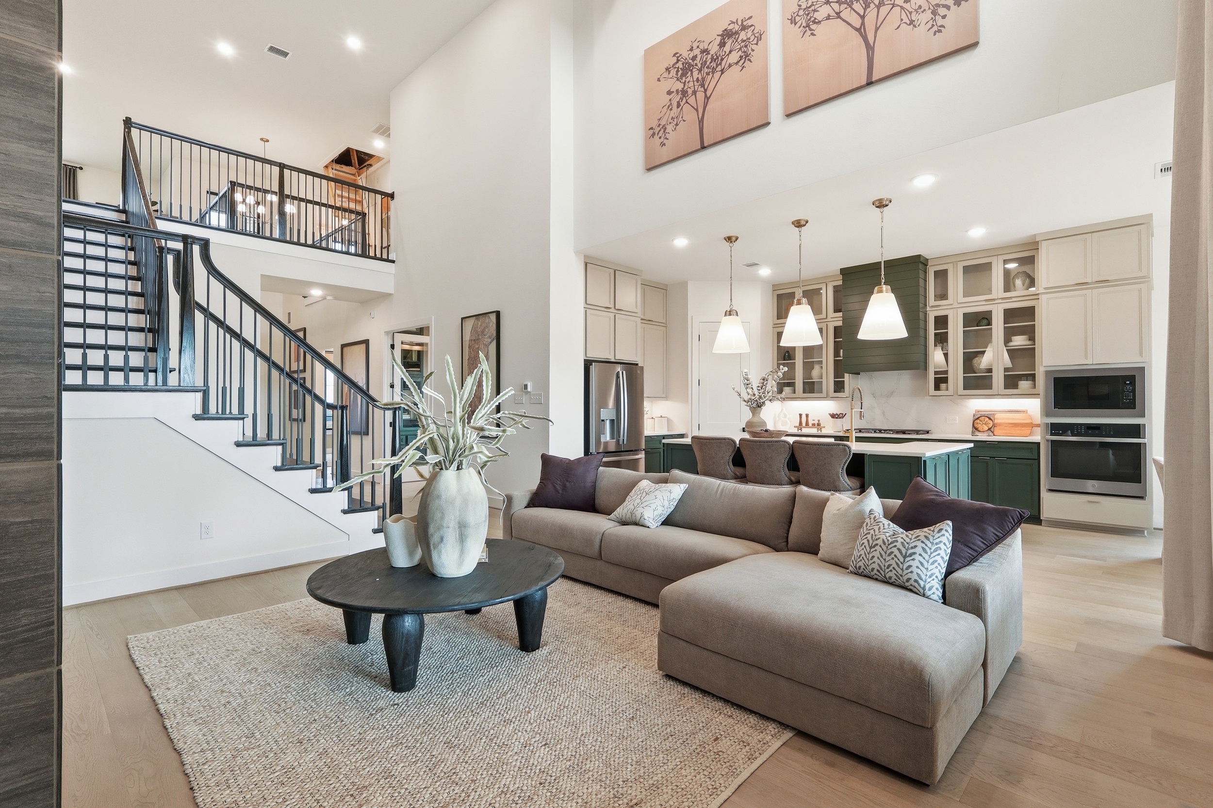 Modern open-concept living room and kitchen with a beige sectional sofa, black coffee table, pendant lights, green lower kitchen cabinets, and a staircase with a black railing leading to an upstairs loft balcony.