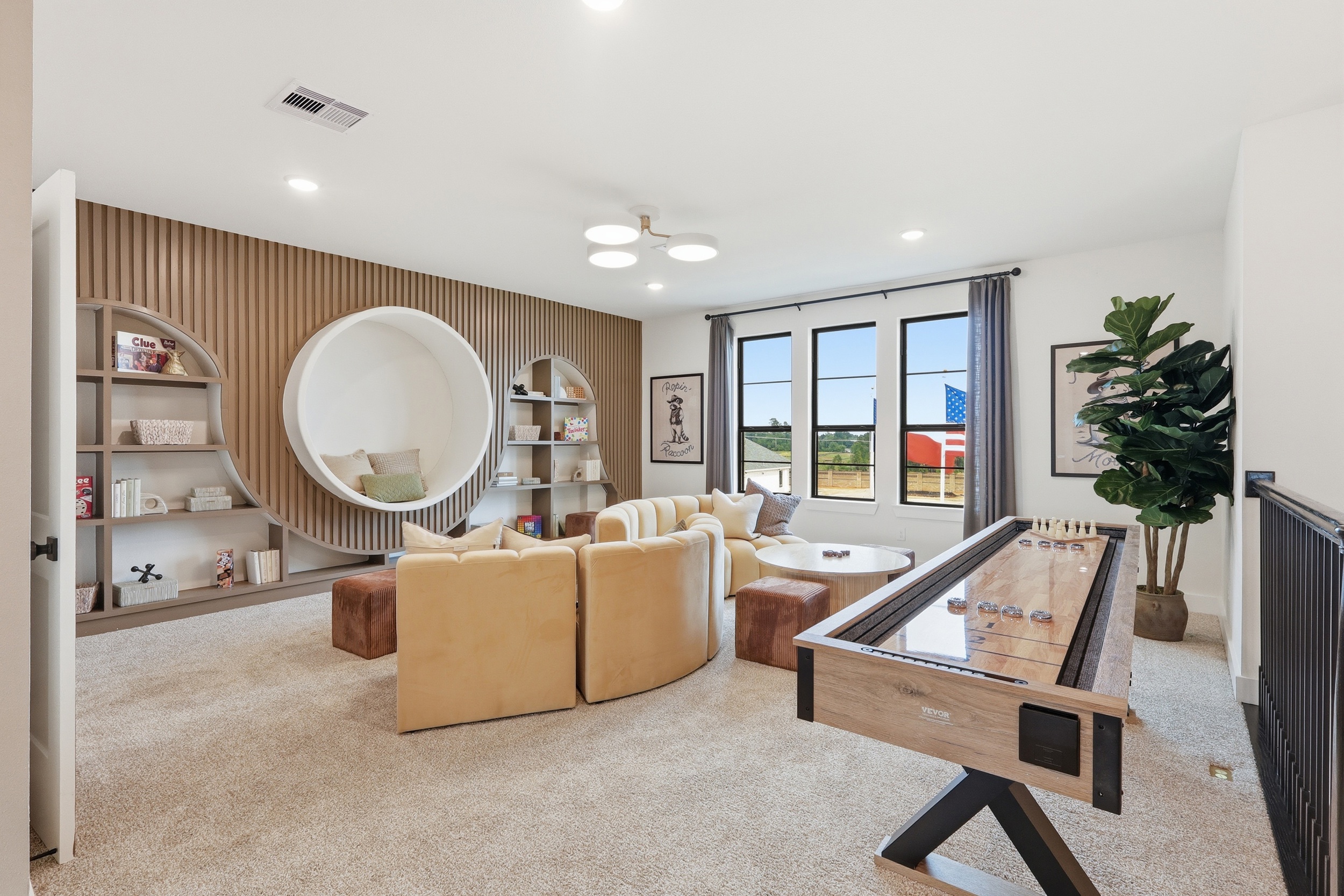 A modern game room with beige carpet, circular shelving, a curved sectional sofa, wall art, a large plant, and a shuffleboard table. Three windows allow natural light into the space.