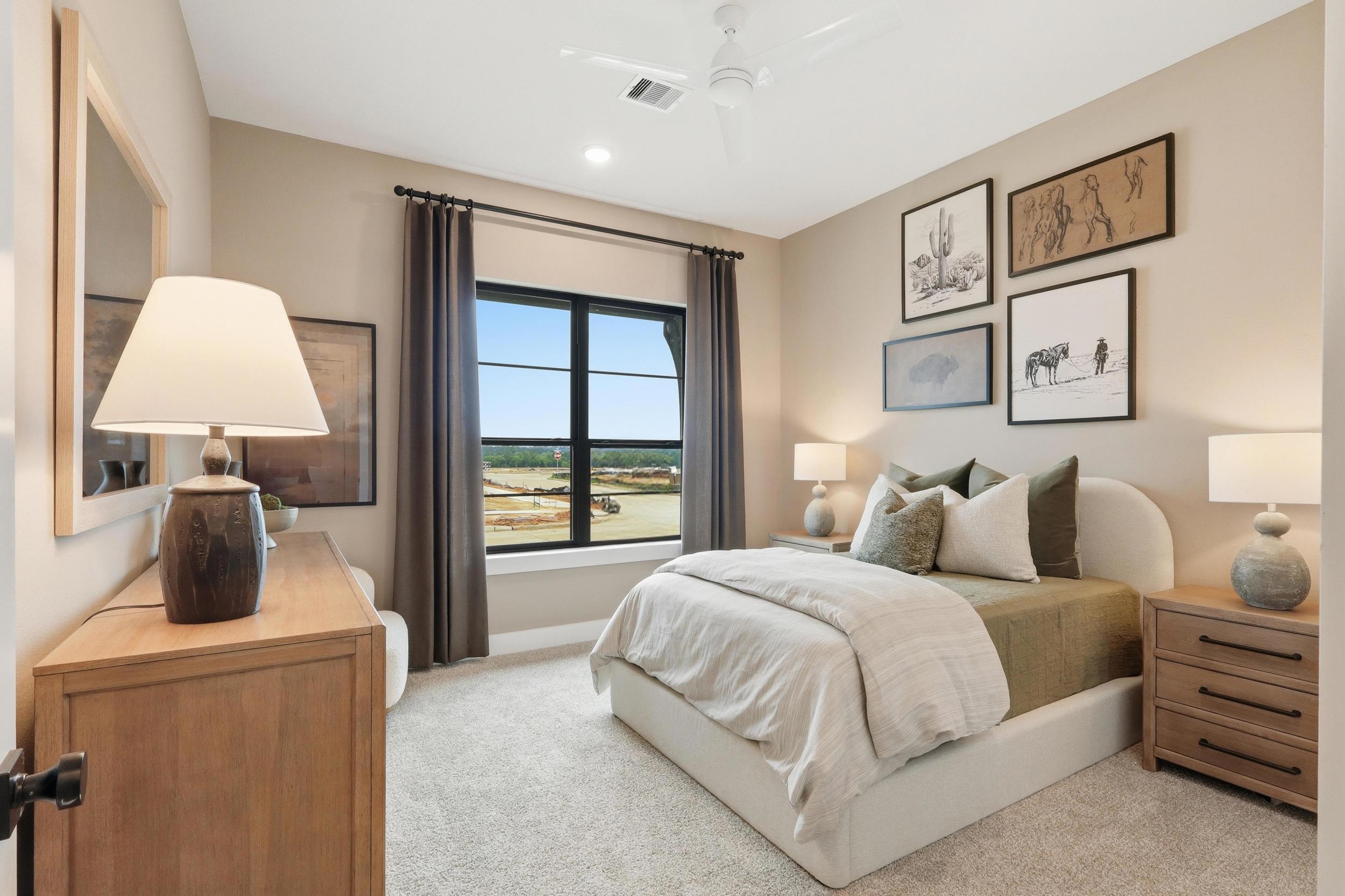 Modern bedroom with a large window, beige walls, neutral bedding, two wooden nightstands, lamps, framed wall art above the bed, and a dresser with decorative items. Sunlight fills the cozy, carpeted space.