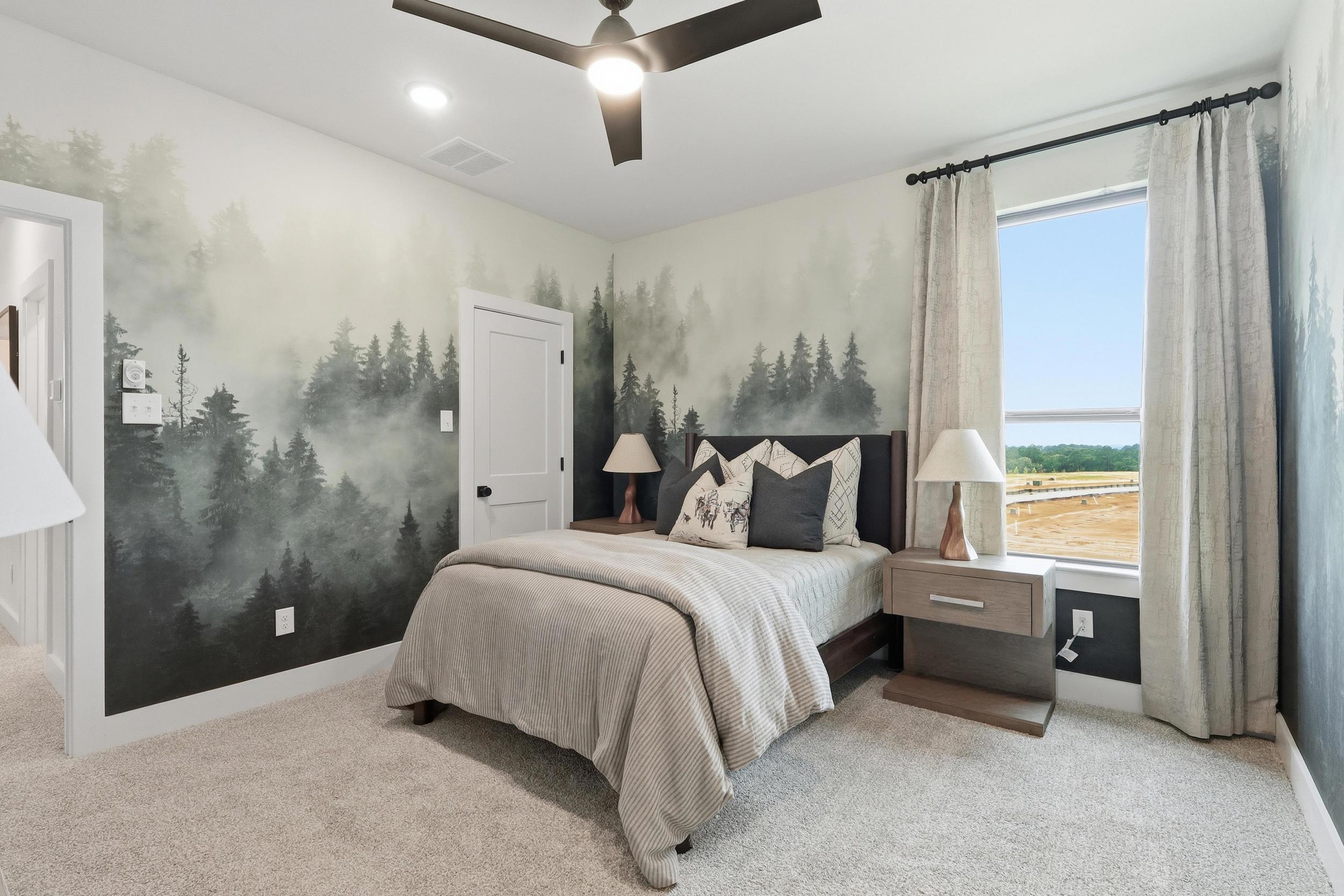 A cozy bedroom with a bed, two nightstands, and lamps. The walls have a misty forest mural, and a large window with curtains lets in natural light and offers a view of a field and water.