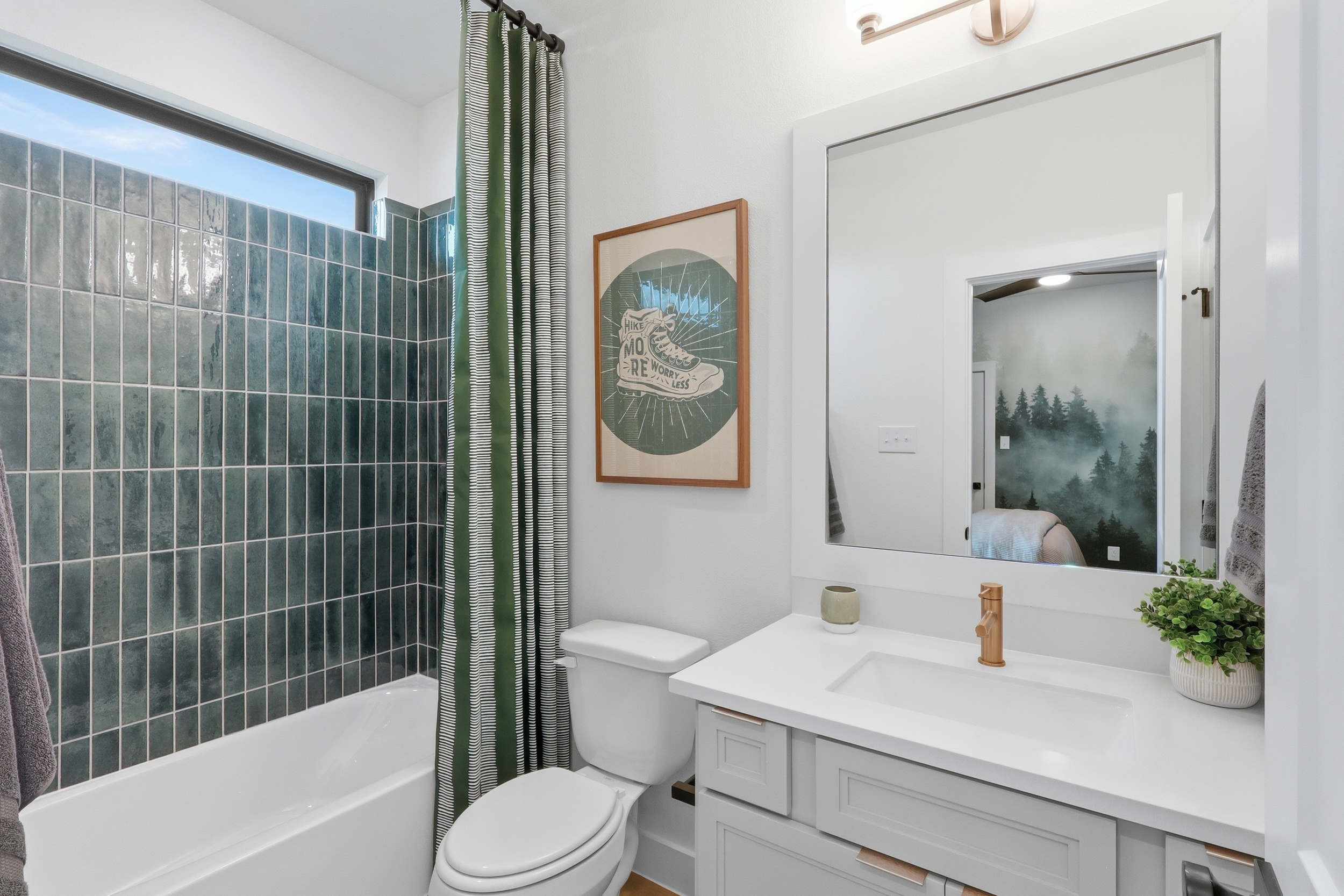 Modern bathroom with green tiled shower, white bathtub, toilet, and white vanity with a sink. A framed sneaker artwork hangs above the toilet, and a green plant sits on the counter next to a large mirror.