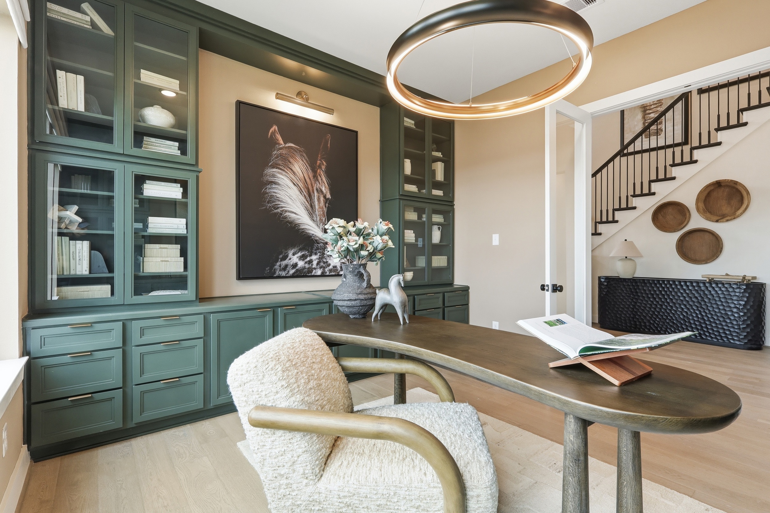 A modern home office with green built-in shelves, a large art piece featuring a horse, a curved desk with an open book, a vase of flowers, and a contemporary light fixture. A staircase is visible through an open doorway.