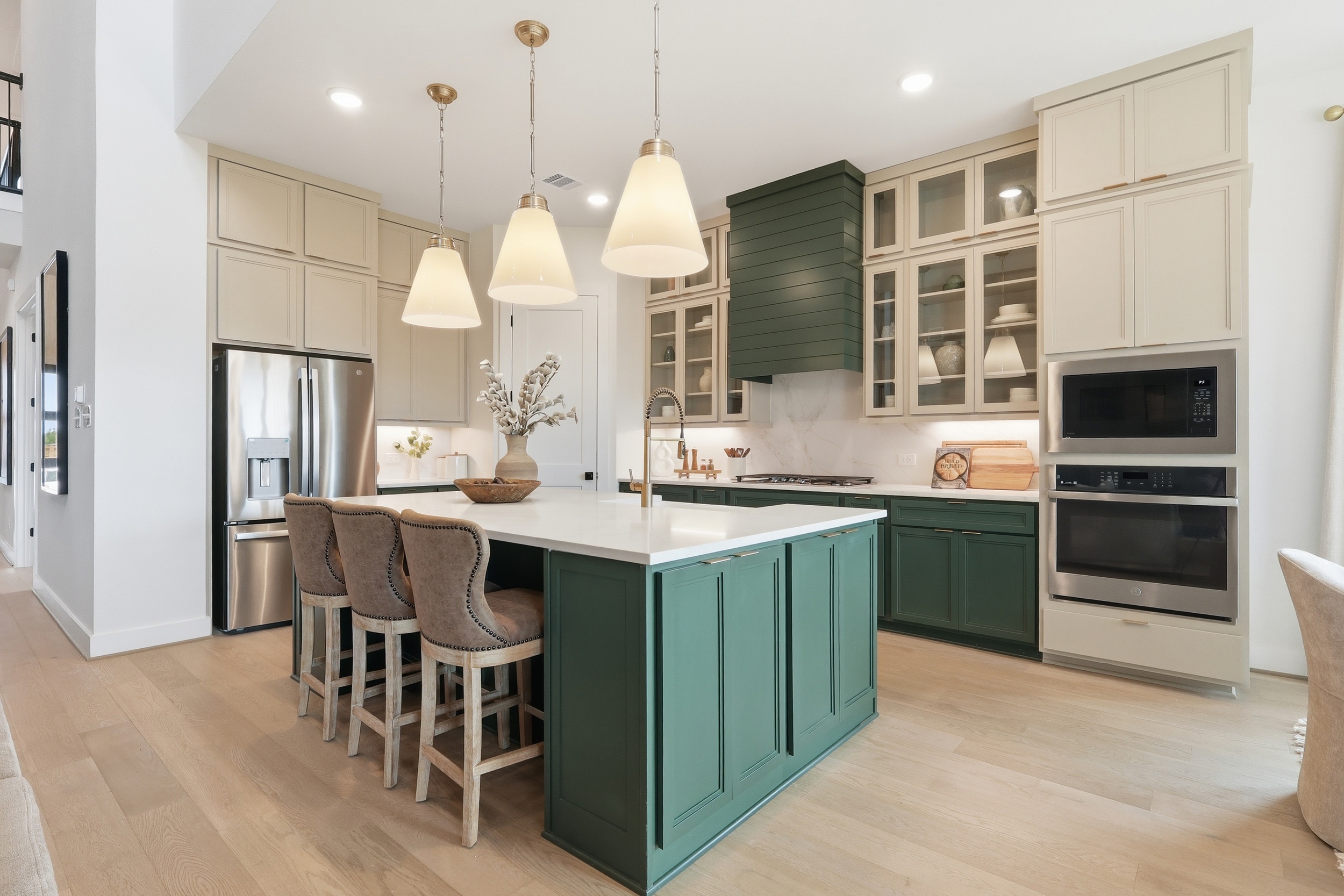 Modern kitchen with green island, beige cabinets, pendant lights, stainless steel appliances, three upholstered bar stools, and light wood flooring. Decorative bowls and branches sit on the island countertop.