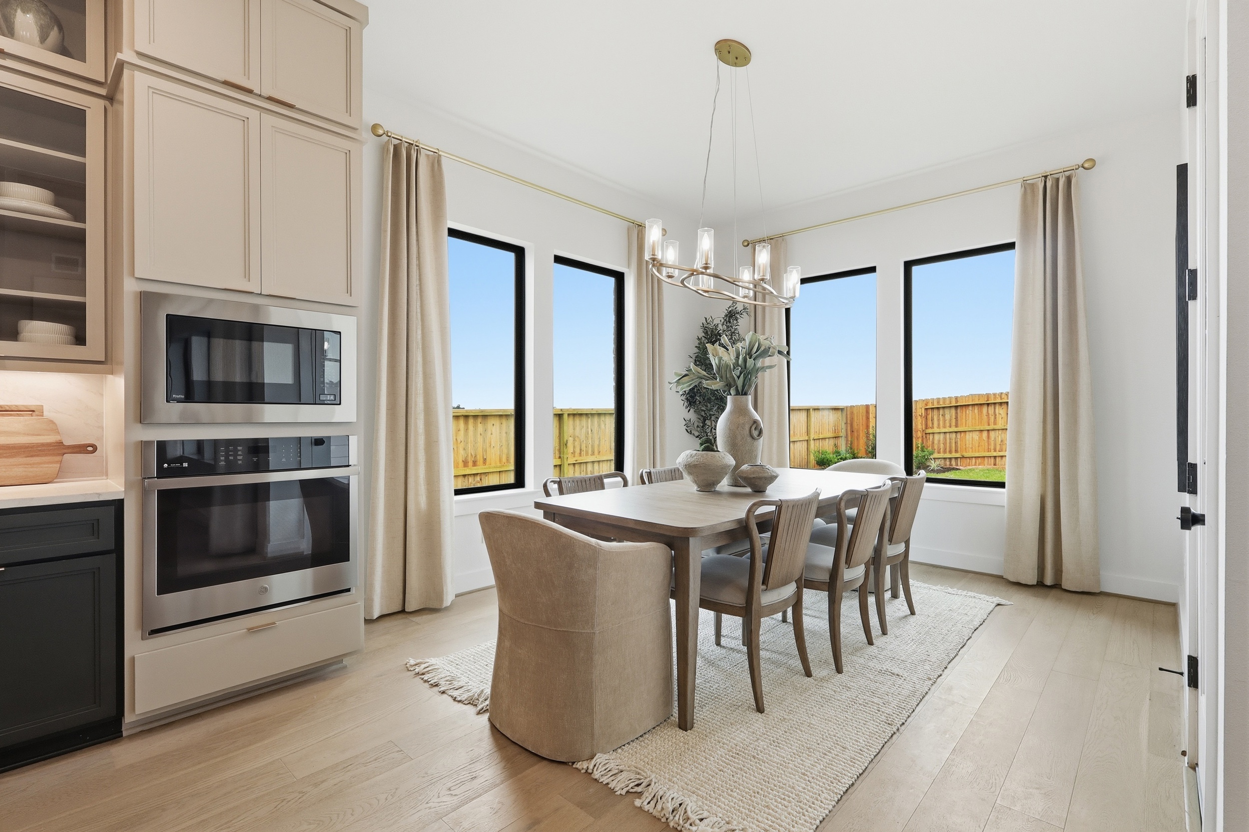 A modern dining area with a wooden table, six chairs, a light chandelier, large windows, beige curtains, and built-in kitchen cabinets with double ovens. The space is bright with natural light and a view of a fenced yard.