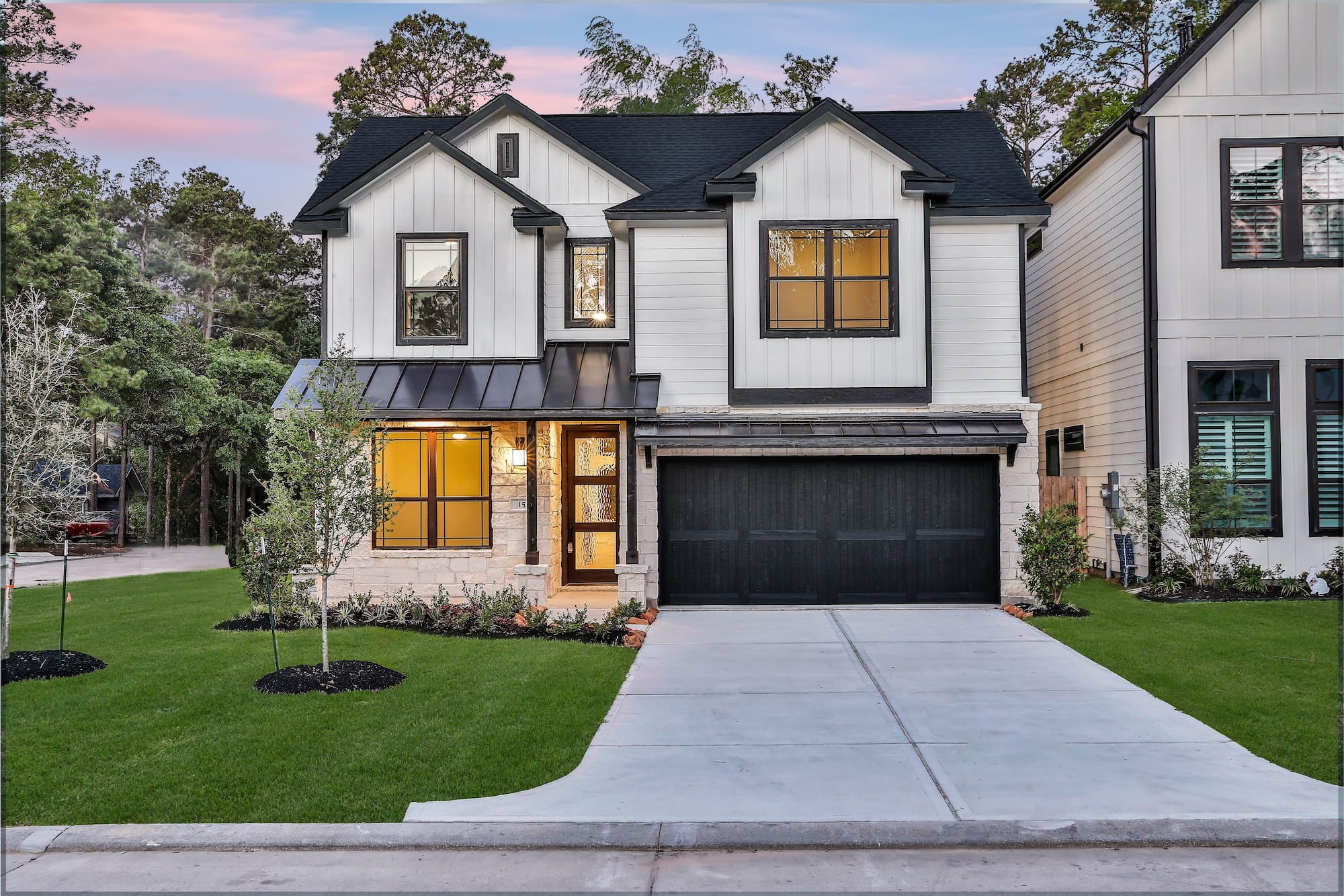 Modern two-story house with white siding, black trim, and a black garage door, featuring a well-kept lawn, small trees, and a sunset sky in the background.