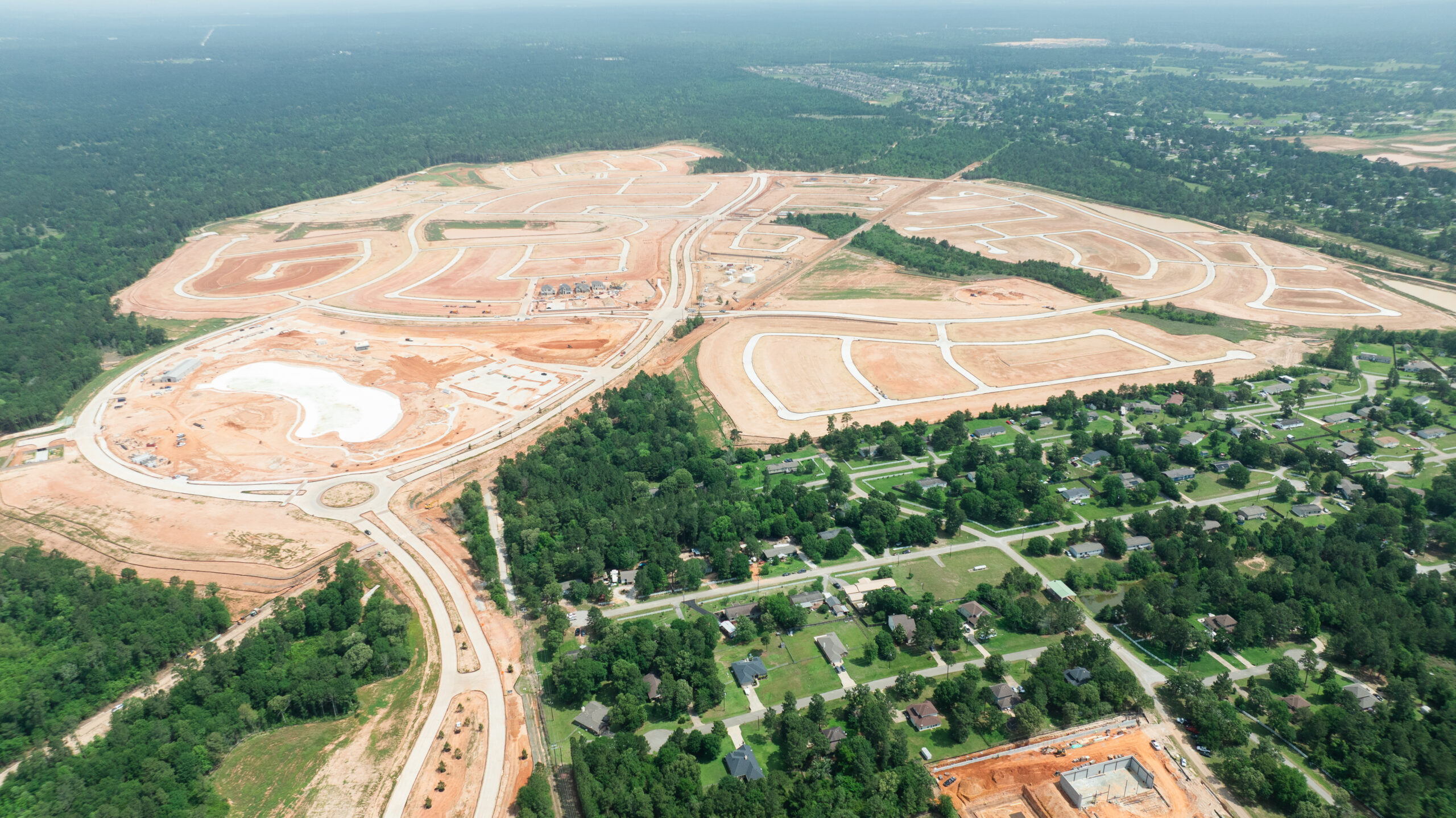 Aerial view of Grand magnolia community in Magnolia, TX under construction, with roads, roundabout, cleared land, and a central lagoon.