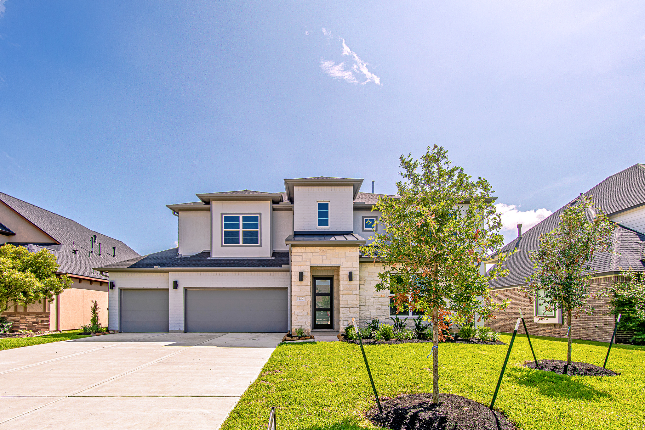 A modern two-story suburban house with a three-car garage, stone and stucco exterior, large driveway, neatly trimmed lawn, young trees, and a clear blue sky overhead.