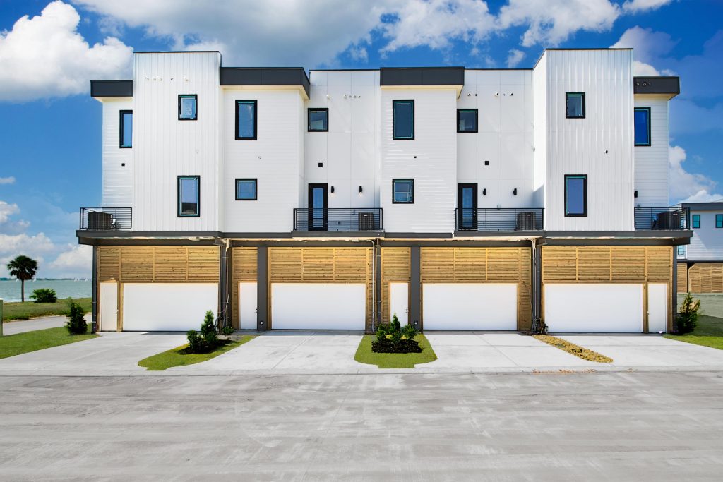 Modern white three-story townhouses with black trim, each featuring a balcony and a two-car garage, set against a blue sky with a few clouds and some landscaping out front.