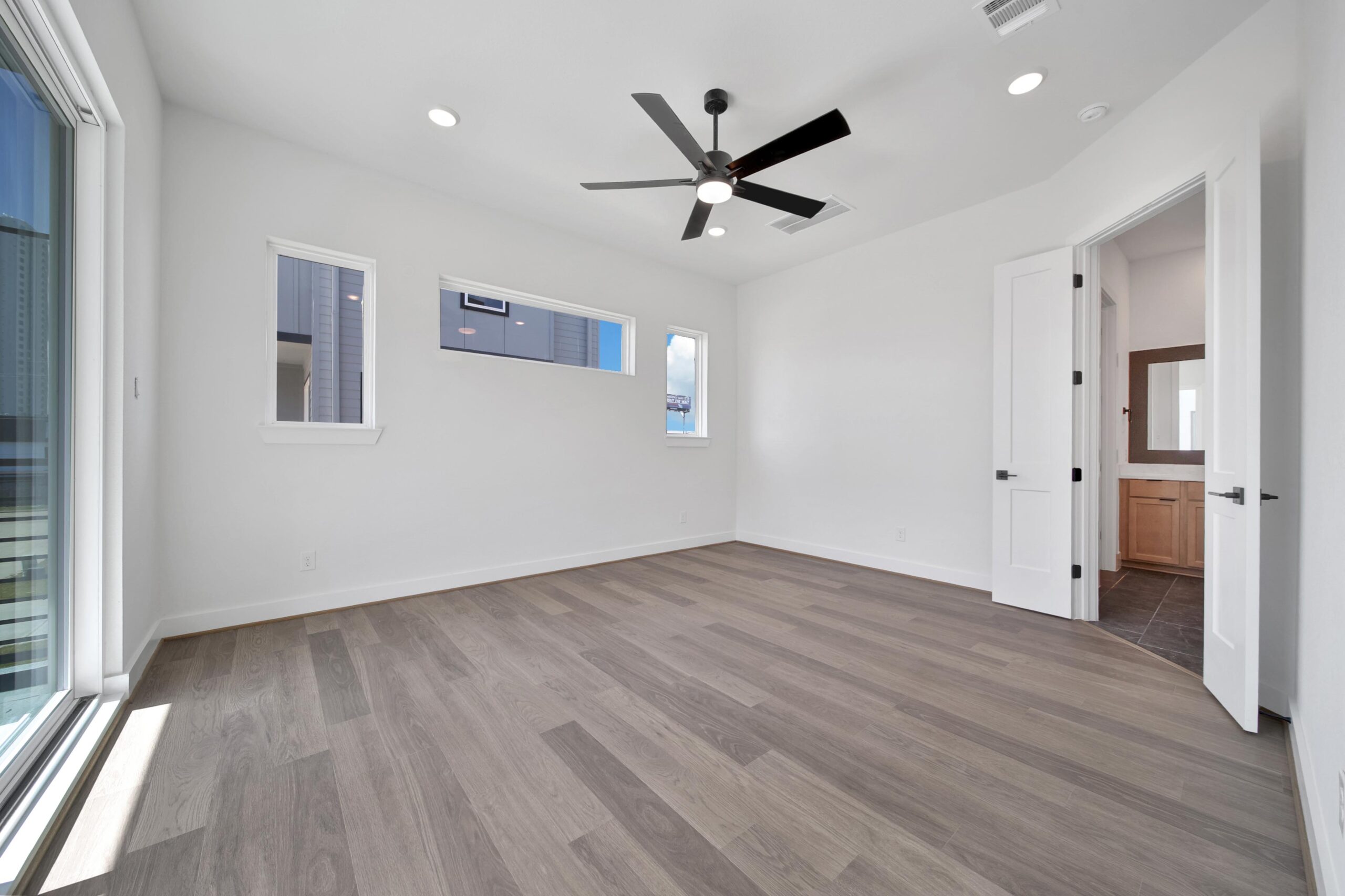 A bright, empty room with light wood flooring, white walls, a black ceiling fan, and several windows. An open door leads to a bathroom with a wood vanity and dark tile floor. Sliding glass doors are on one side.
