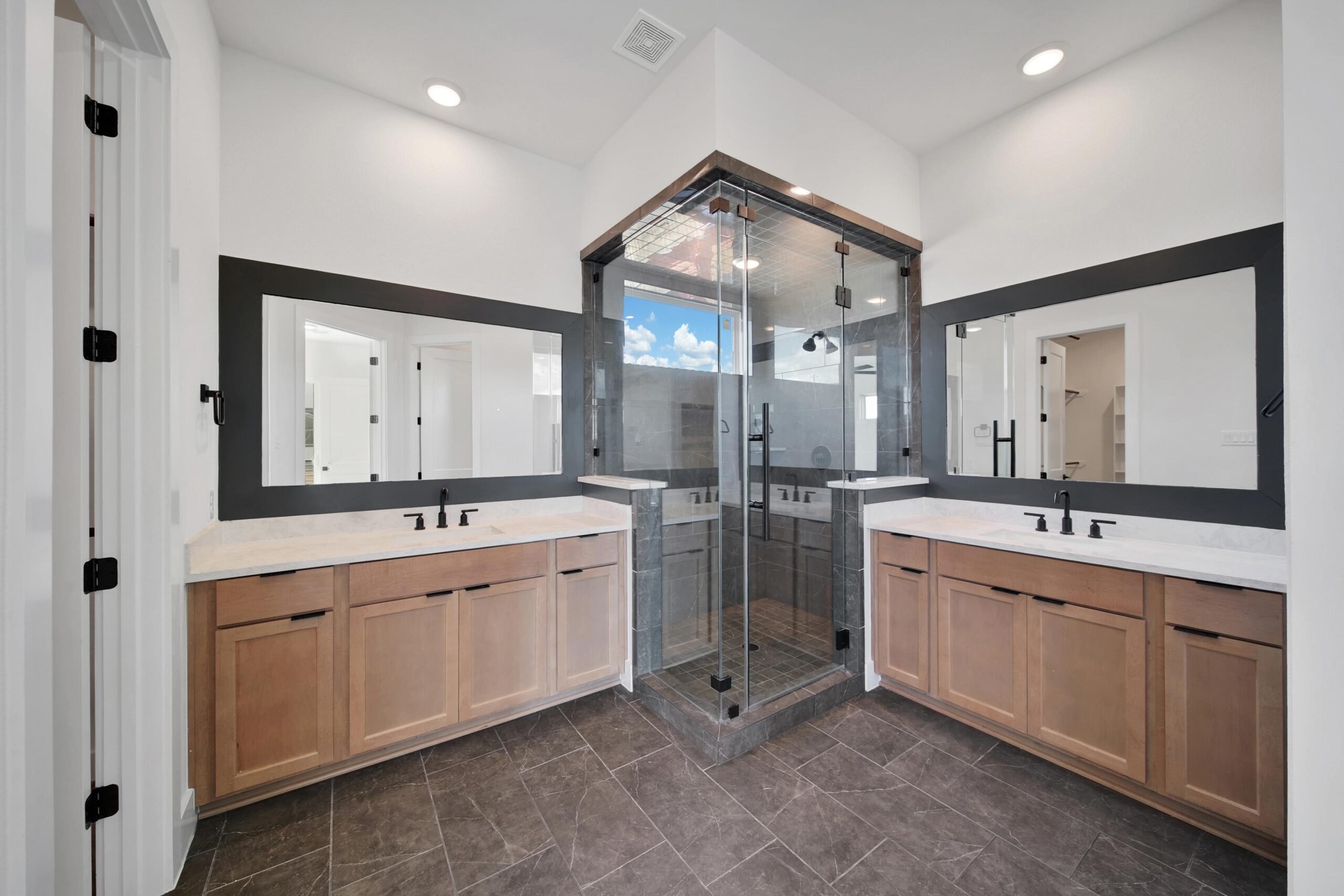 Modern bathroom with double vanities on each side, light wood cabinets, black fixtures, and a large glass-enclosed shower in the center; mirrors and recessed lighting on white walls.