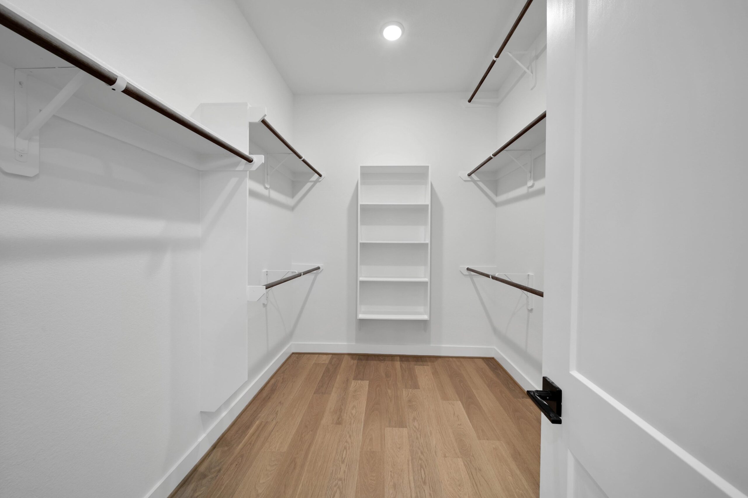 Empty walk-in closet with white walls, wooden floor, open door, built-in shelves at the far end, and multiple white shelves with wooden hanging rods on both sides. Ceiling light illuminates the space.