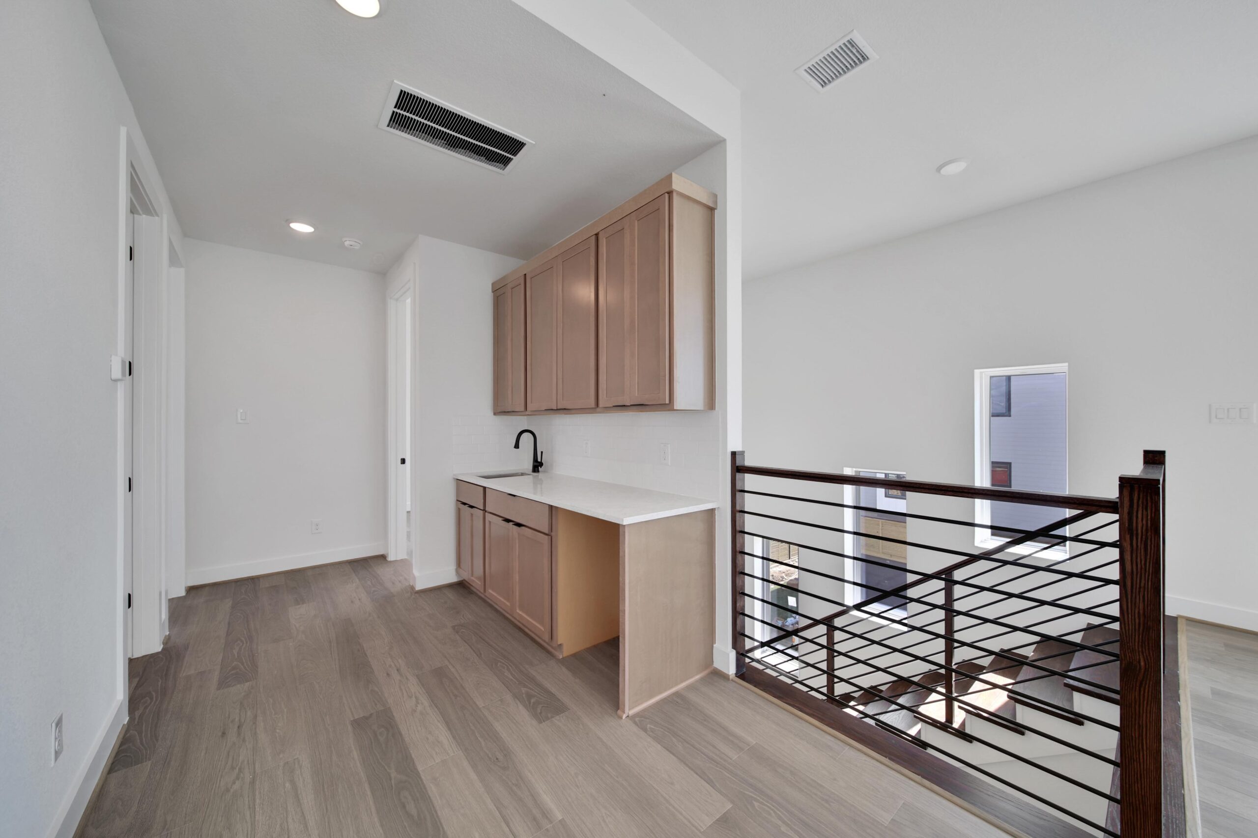 A modern hallway with light wood flooring, built-in cabinets and countertop with a sink, and a black railing overlooking the floor below. White walls and natural light create a bright, open feel.
