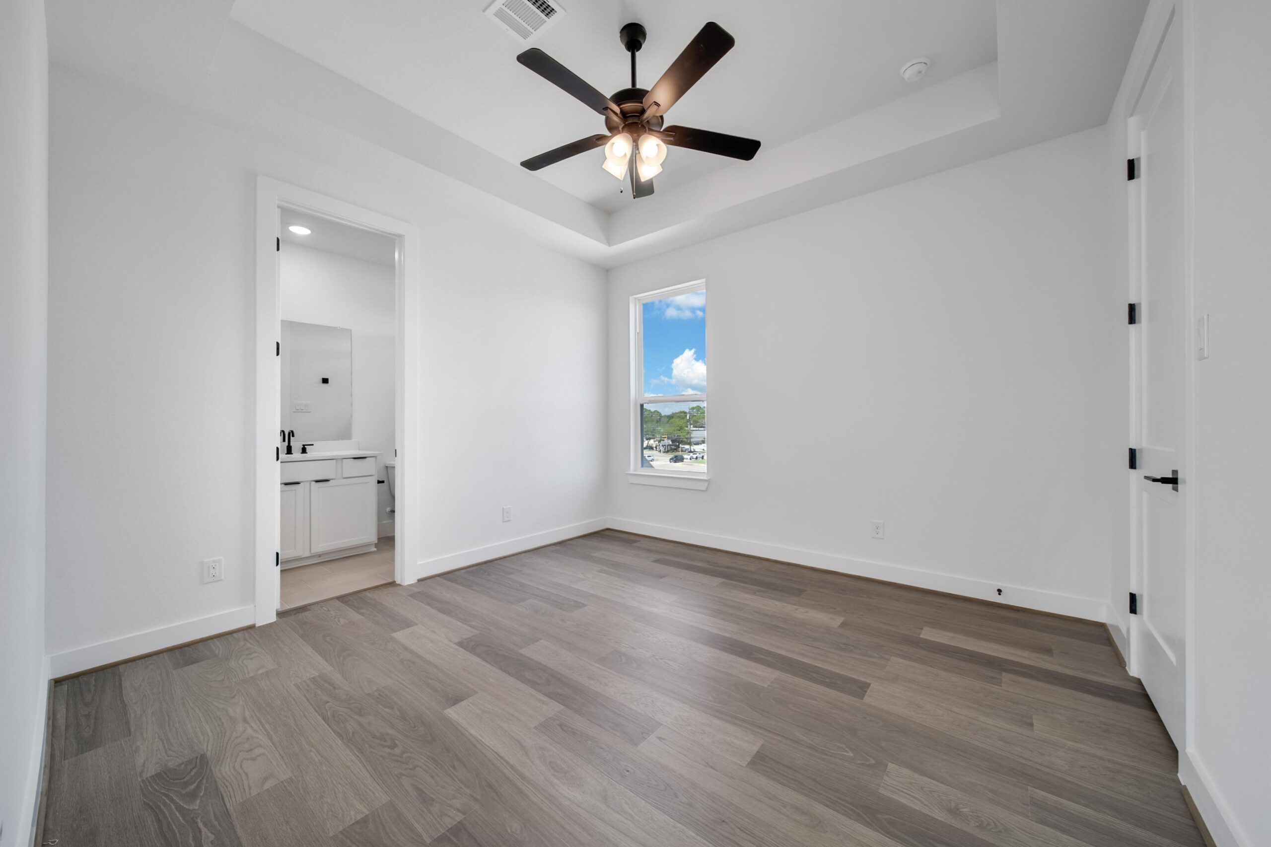 Empty modern bedroom with wood flooring, white walls, a ceiling fan with lights, a window showing a blue sky and greenery, and an open door leading to a bathroom with a sink and mirror.