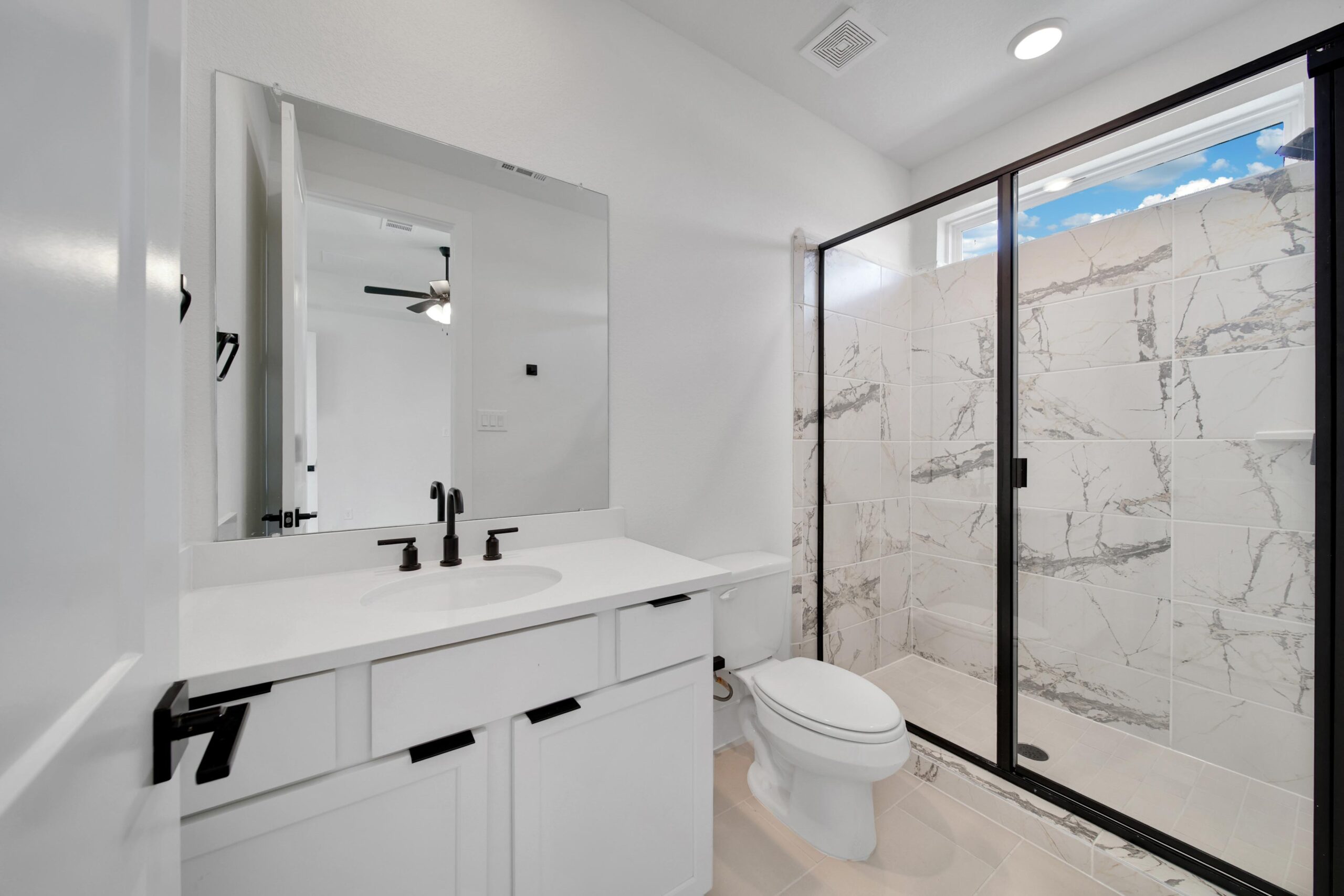 Modern bathroom with white walls, a vanity with black fixtures, a toilet, and a large walk-in shower featuring glass doors and white marble-patterned tiles. A mirror reflects part of an adjacent room.