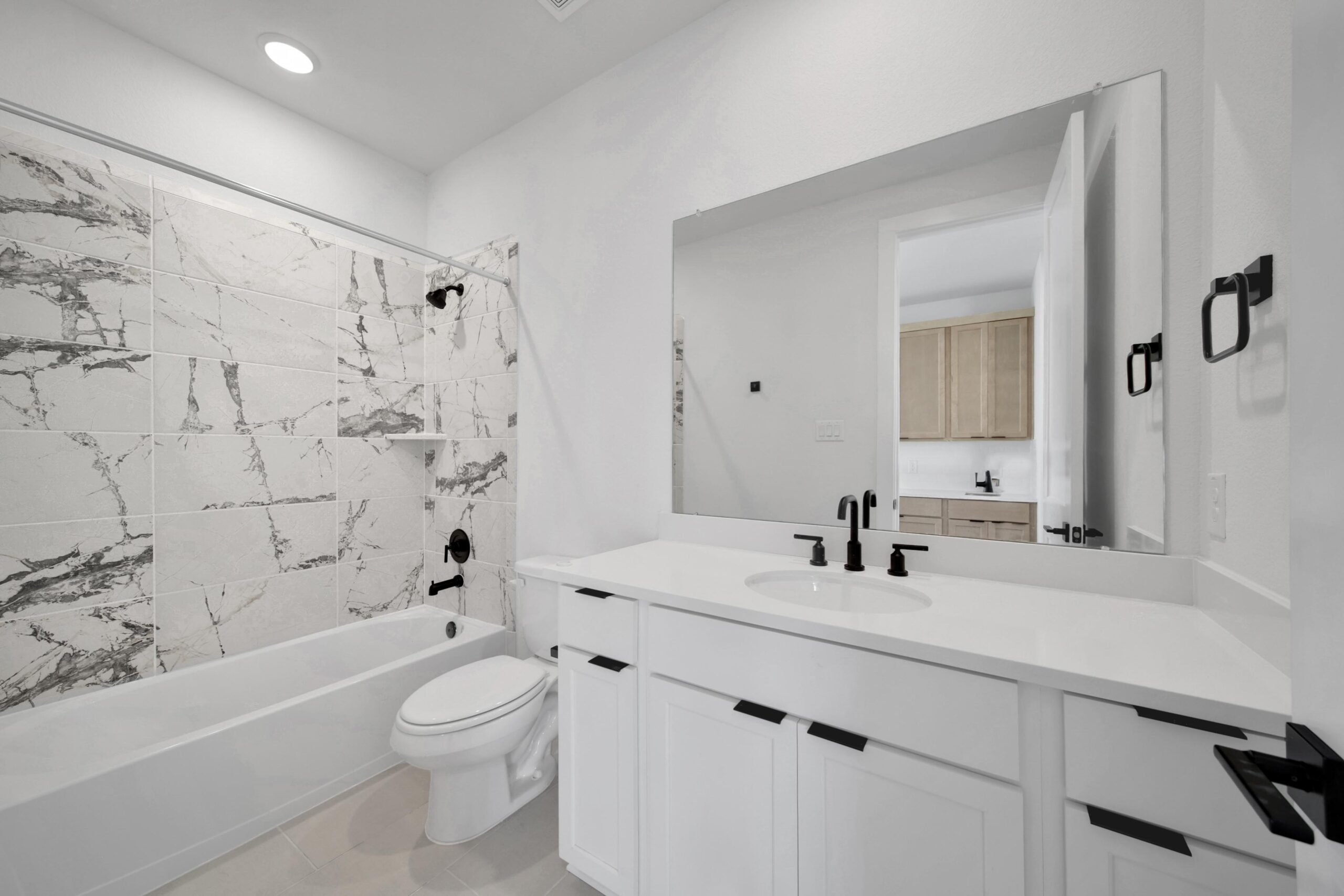 Modern bathroom with white cabinets, large mirror, white countertop, and black fixtures. Bathtub with marbled tile surround and black showerhead. Toilet beside the tub. Open door reveals a glimpse of another room.