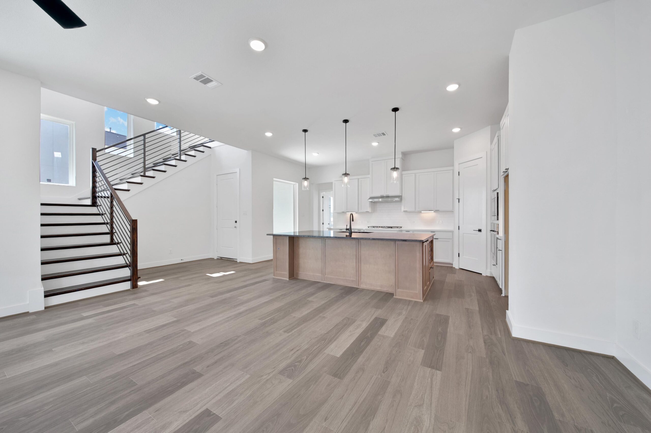 Modern kitchen with a large island, white cabinets, stainless steel appliances, pendant lights, and light wood flooring. Open layout includes a staircase with dark railings and a bright, airy atmosphere.