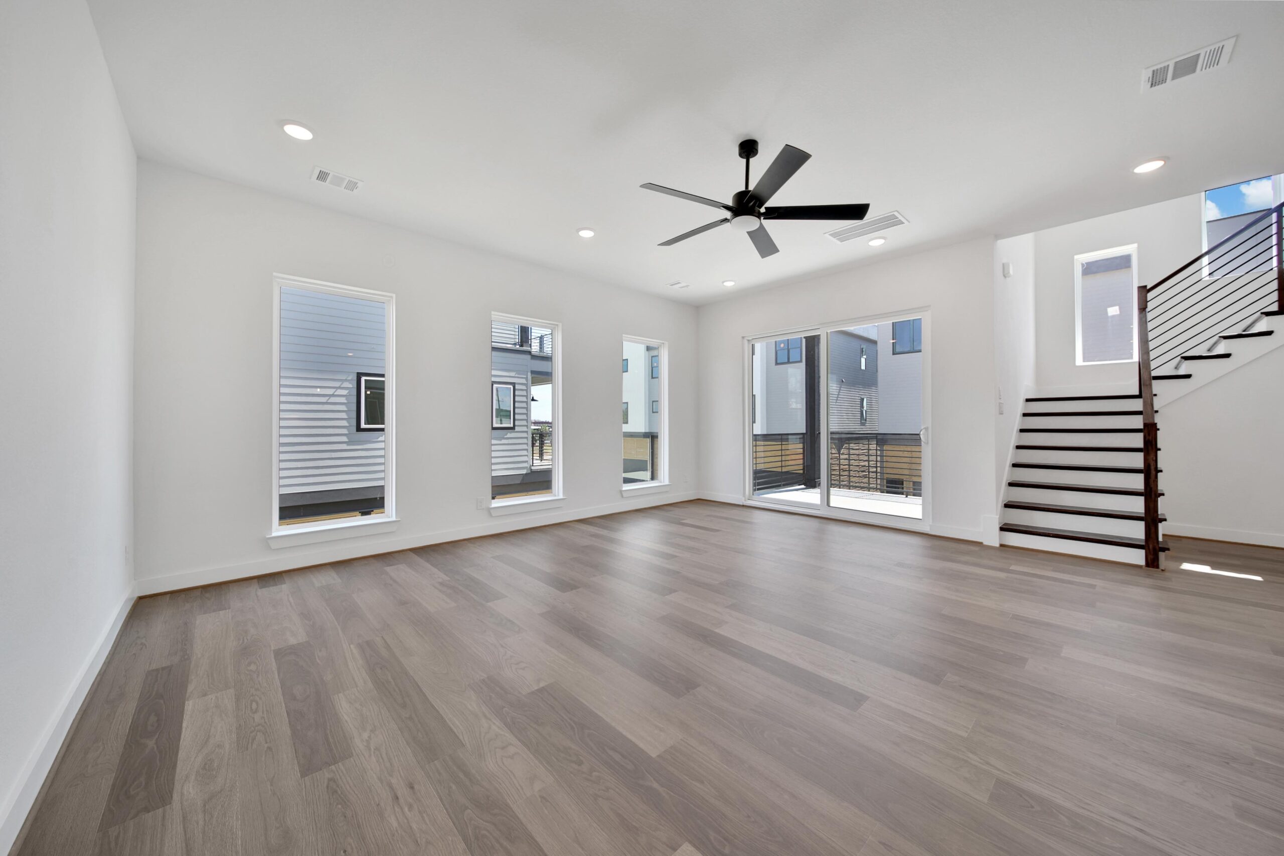 A spacious, modern living room with light wood flooring, white walls, large windows, sliding glass doors, a black ceiling fan, and an open staircase leading to the upper floor.