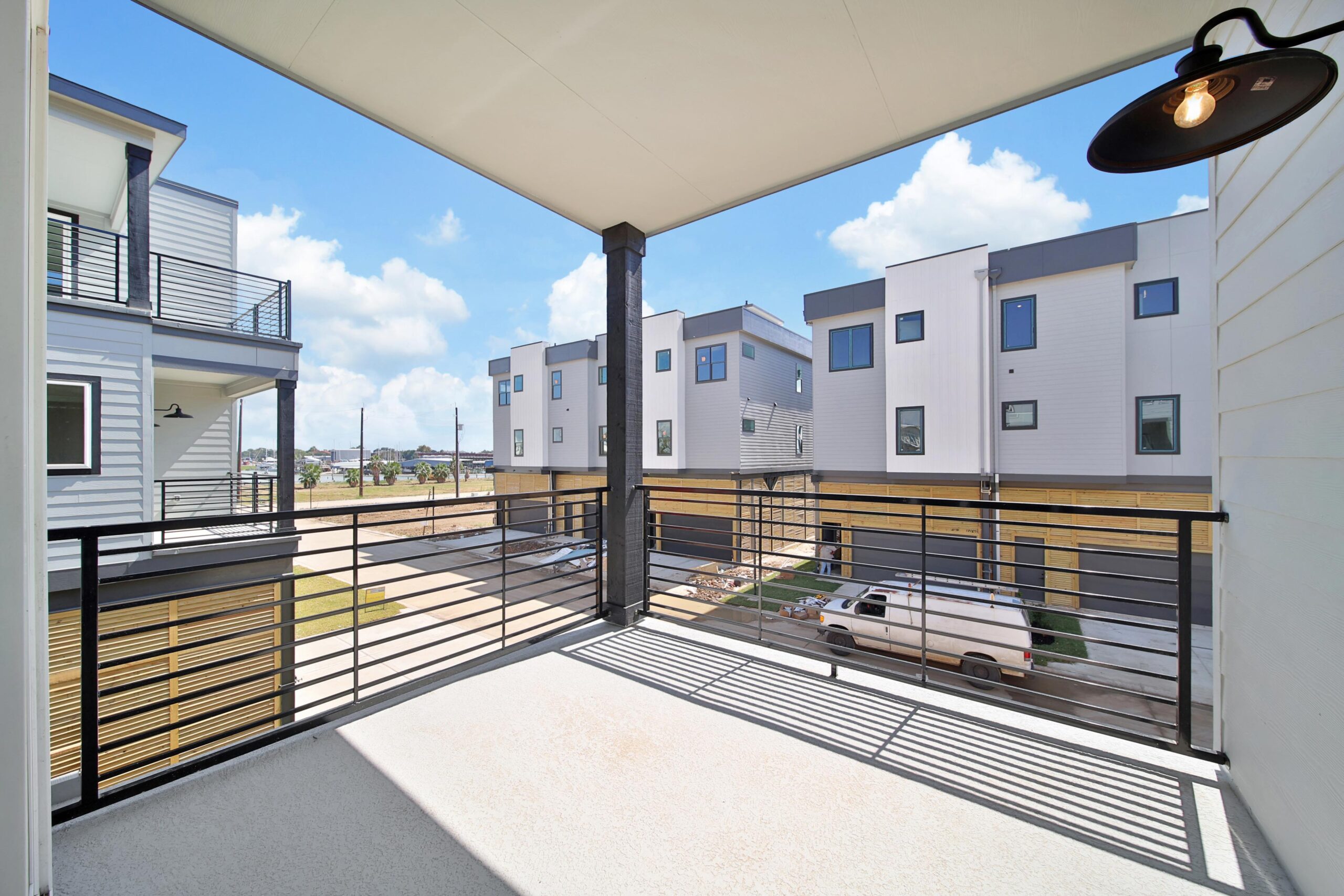 View from a covered balcony with a black metal railing, overlooking a residential area with modern, multi-story white and gray townhouses and a parked white pickup truck, under a bright blue sky with scattered clouds.