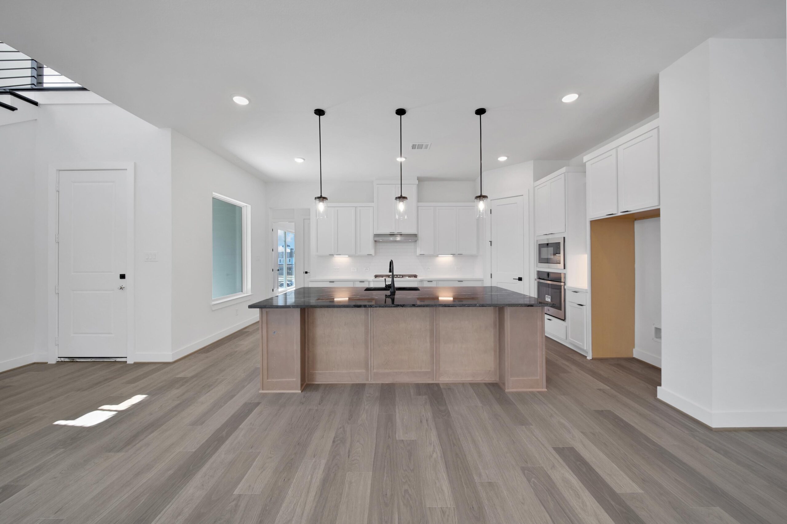 Modern kitchen with light wood flooring, white cabinets, a large island with a dark countertop, three pendant lights above the island, and built-in stainless steel appliances. The space is bright with recessed lighting.