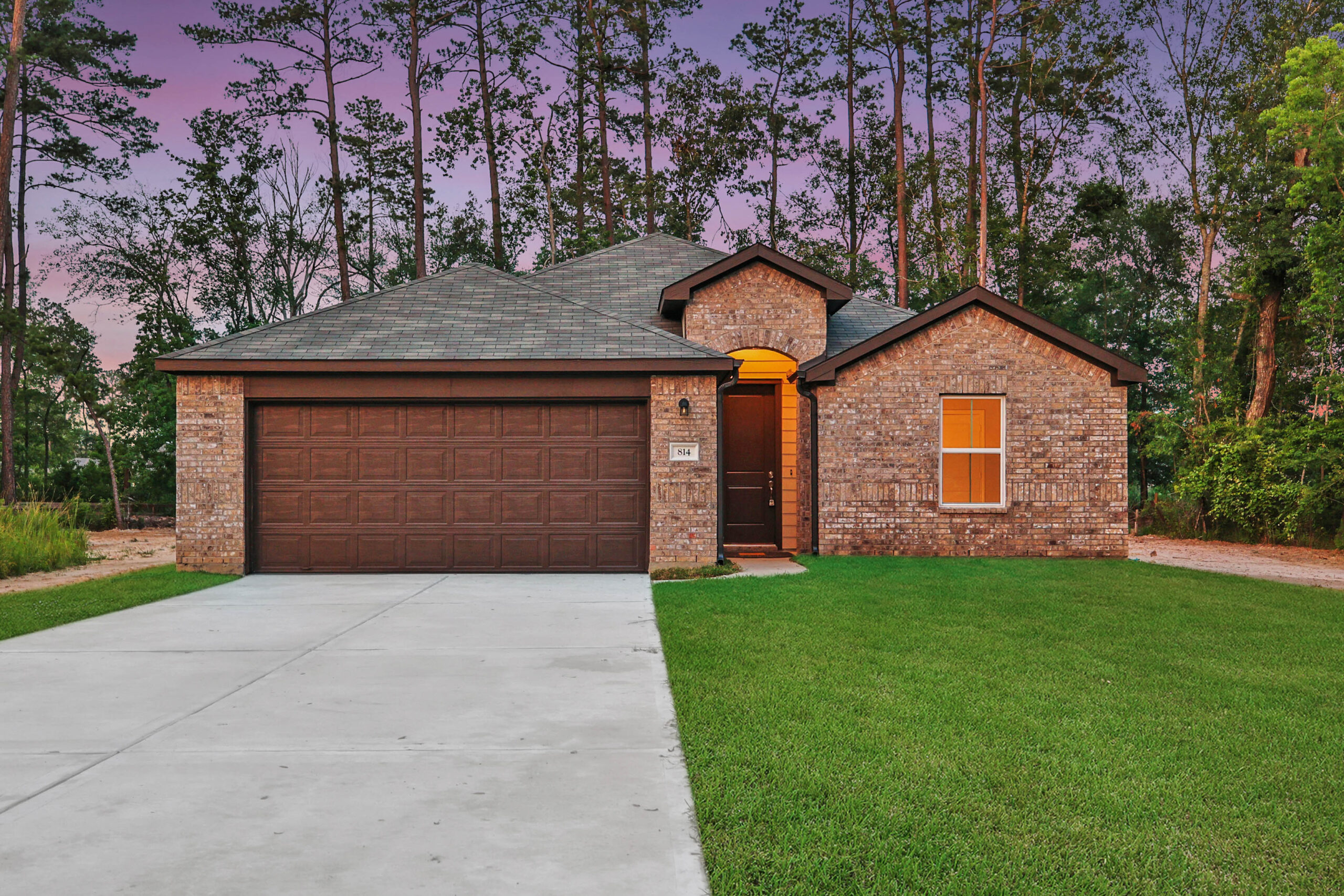 Single-story brick house with a dark brown garage door, well-kept lawn, and a concrete driveway. The home is surrounded by tall trees, and the sky is purple at sunset. Warm lights are visible through the windows and entryway.