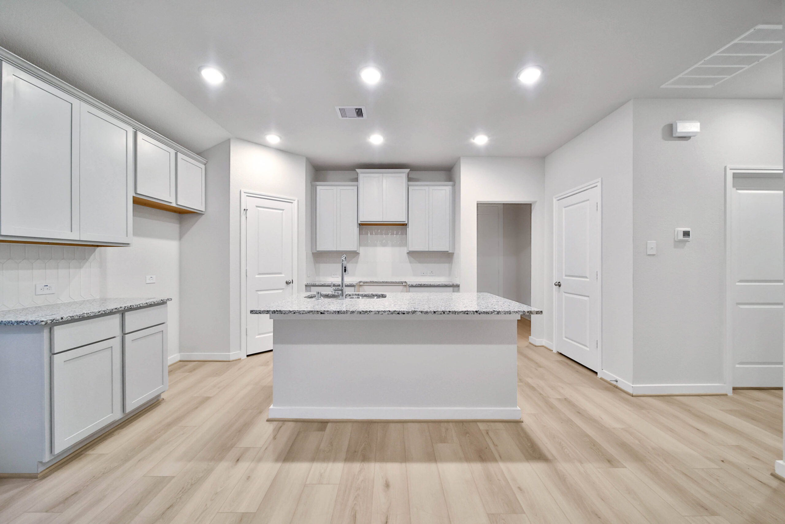 Modern, bright kitchen with white cabinets, granite countertops, an island in the center, light wood flooring, and recessed ceiling lights. There are multiple doors and minimal decor, giving it a clean, spacious look.