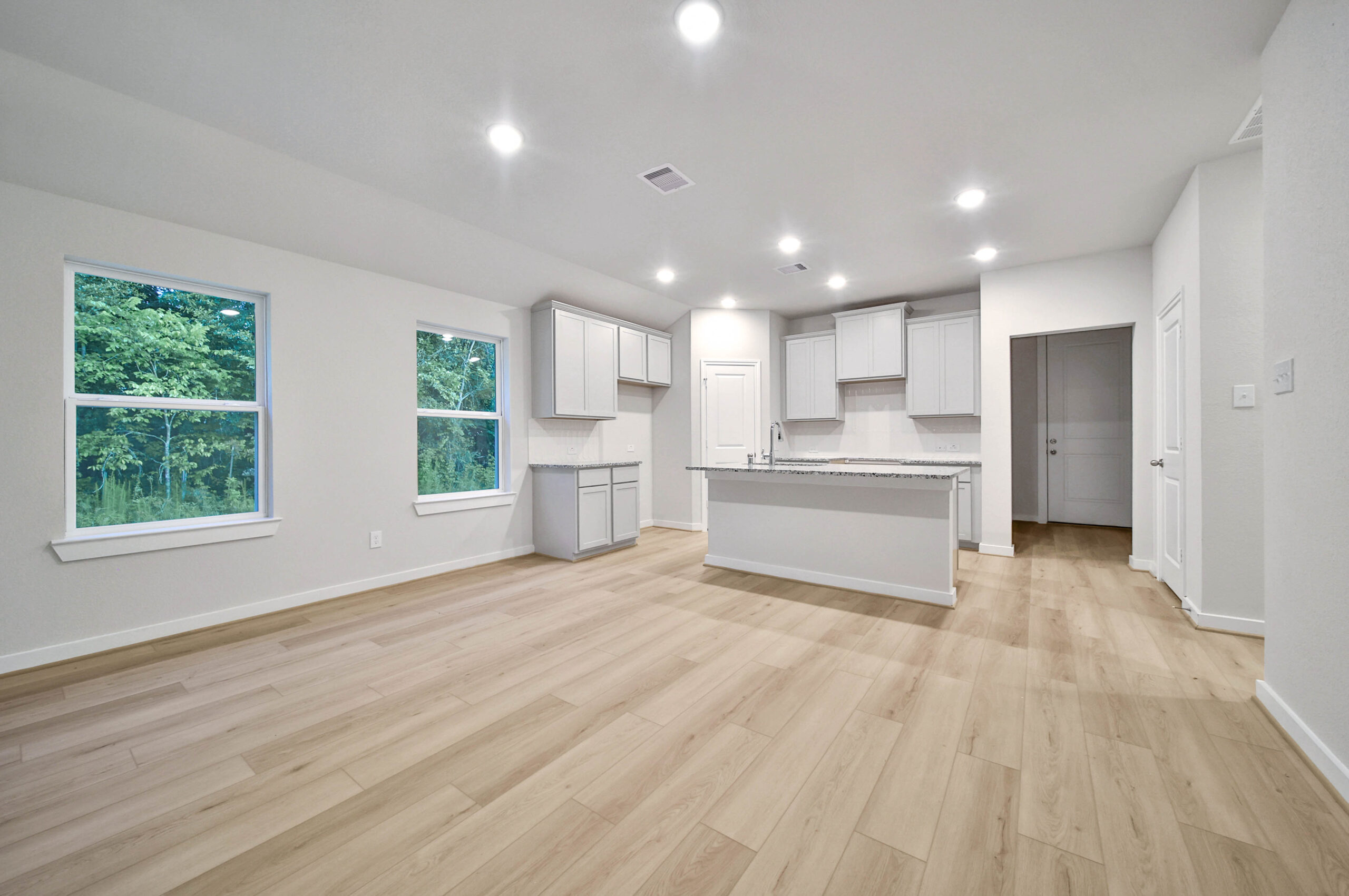Spacious modern kitchen and living area with light wood flooring, white cabinets, large island, and recessed lighting. Two windows allow natural light and provide a view of green trees outside.