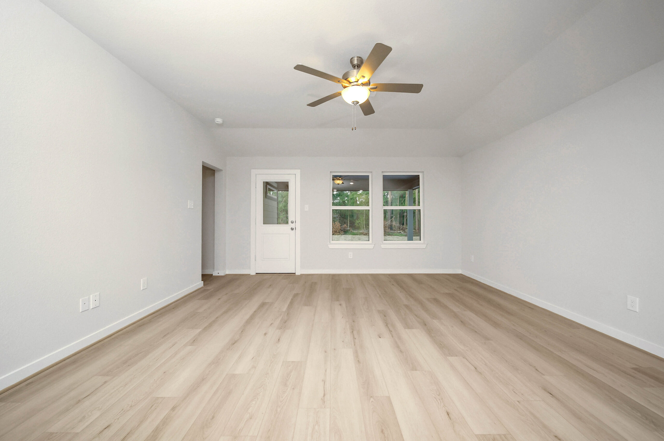 A spacious, empty room with light wood flooring, white walls, a ceiling fan with a light, two windows, and a glass door leading outside.