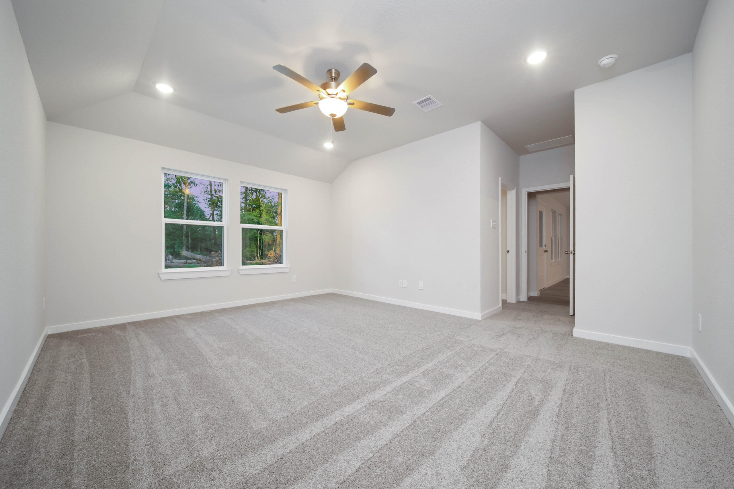 Spacious empty room with light gray carpet, white walls, a ceiling fan with lights, two large windows, and an open doorway leading to a hallway.