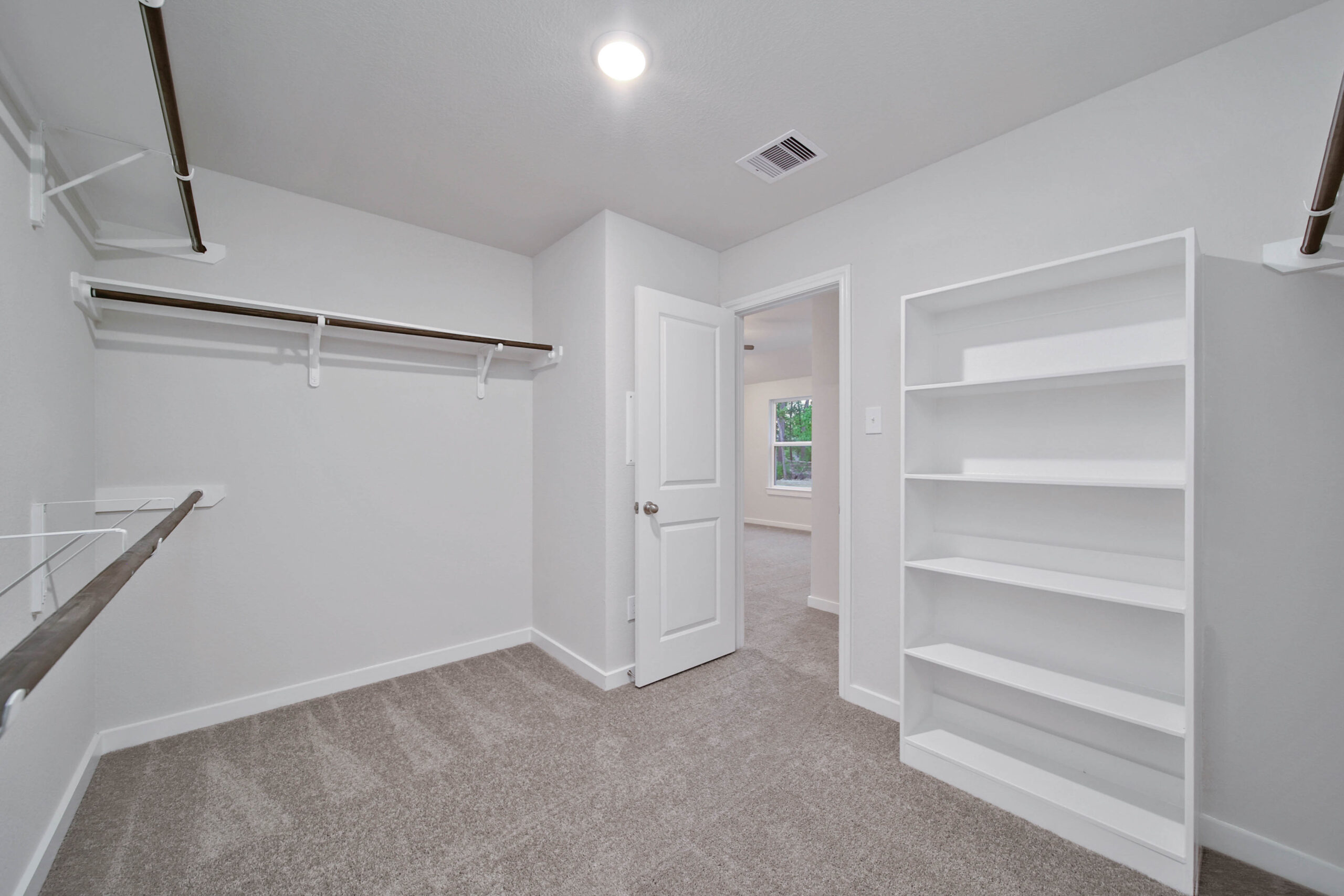 Spacious walk-in closet with white walls, built-in shelves, hanging rods, beige carpet flooring, and a door leading to a bright room with windows. Ceiling light and air vent are visible.