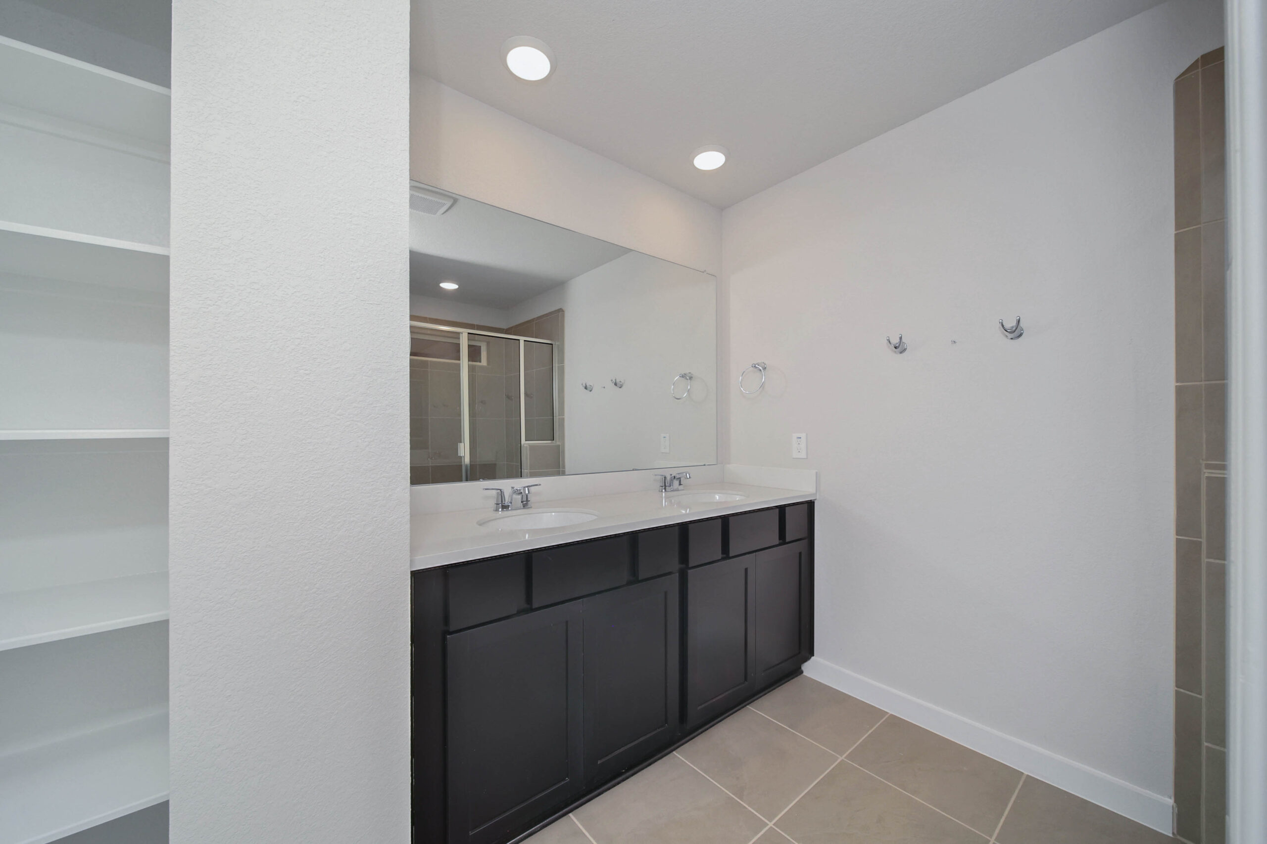 A modern bathroom with a double-sink vanity, large wall mirror, dark cabinets, beige tiled floor, built-in open shelving on the left, and neutral-colored walls. Recessed ceiling lights provide bright illumination.