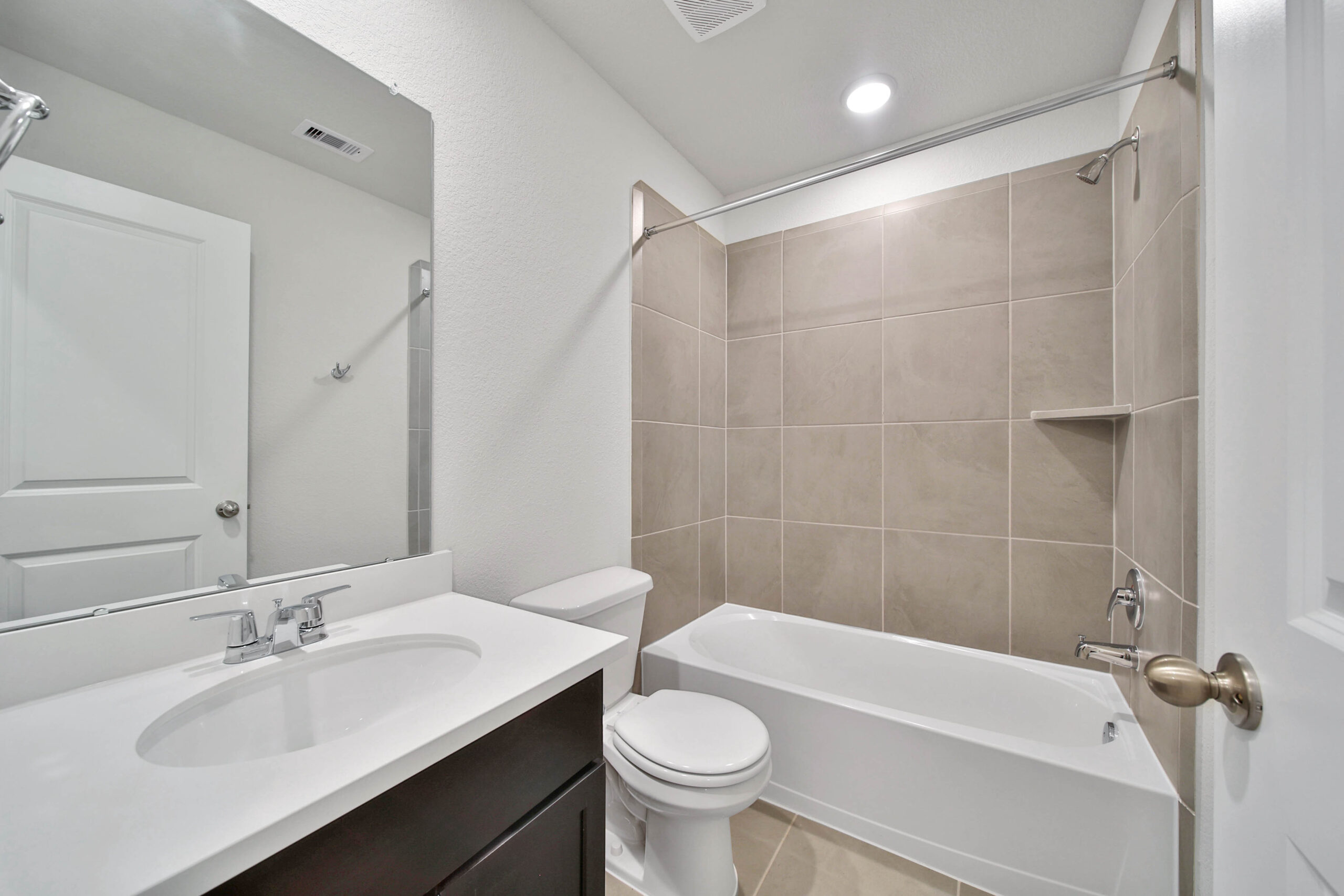 Modern bathroom with a white sink vanity, silver faucet, toilet, and bathtub with beige tile surround. Large mirror above the sink and bright overhead lighting create a clean, minimalist look.