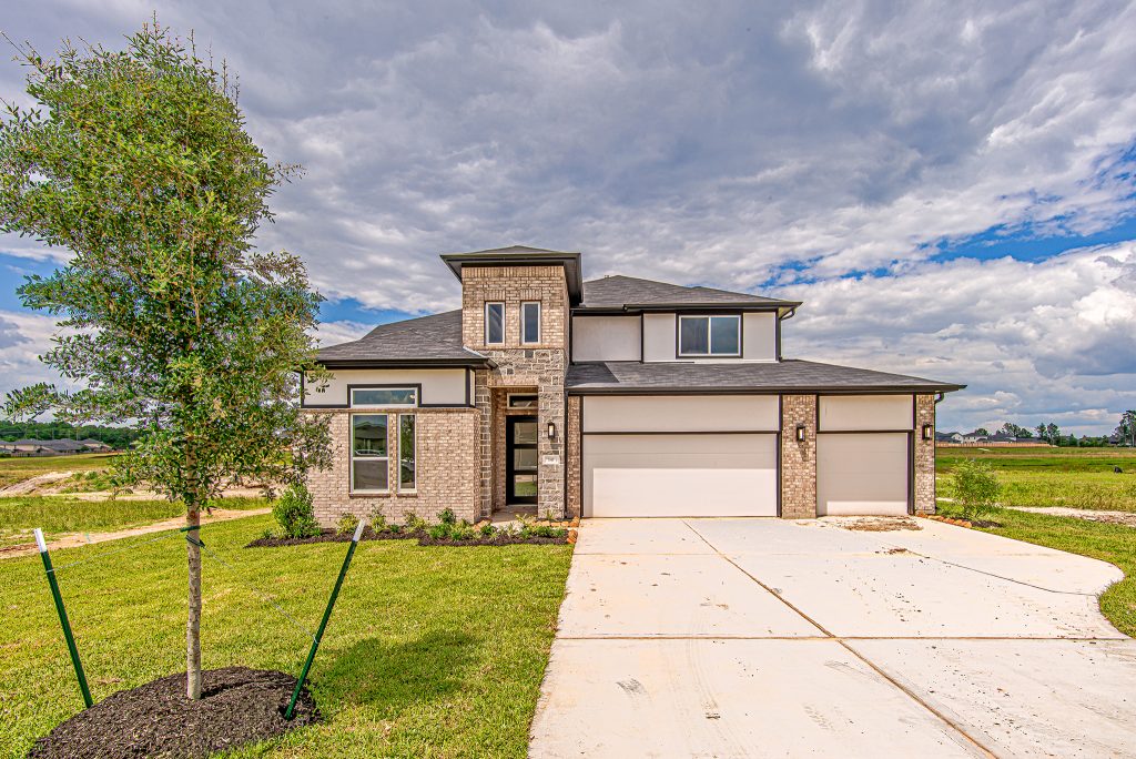 Modern two-story brick house with a three-car garage, large driveway, manicured lawn, and a newly planted tree in the front yard under a partly cloudy sky.