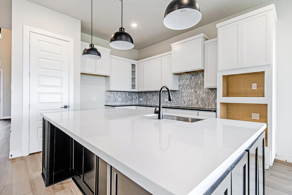 Modern kitchen with white cabinets, a large white island countertop, black fixtures, pendant lights, a mosaic tile backsplash, and light wood flooring. Open shelving and a built-in space are visible on the right.