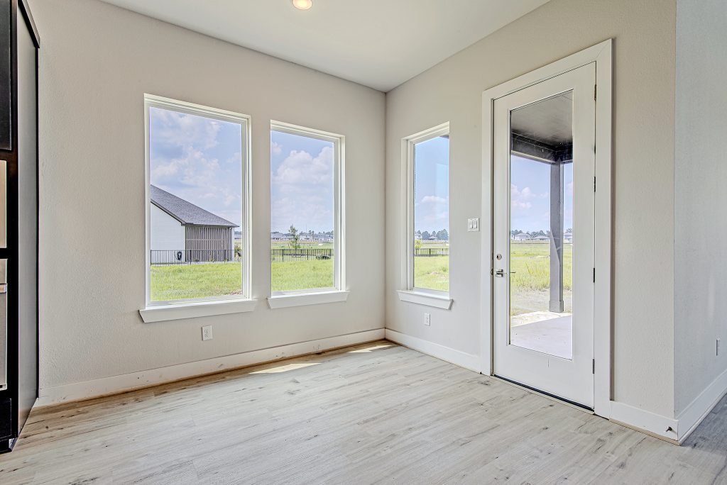 A bright, empty room with light wood flooring, three large windows, and a glass door offering views of a grassy yard and a neighboring house under a partly cloudy sky.