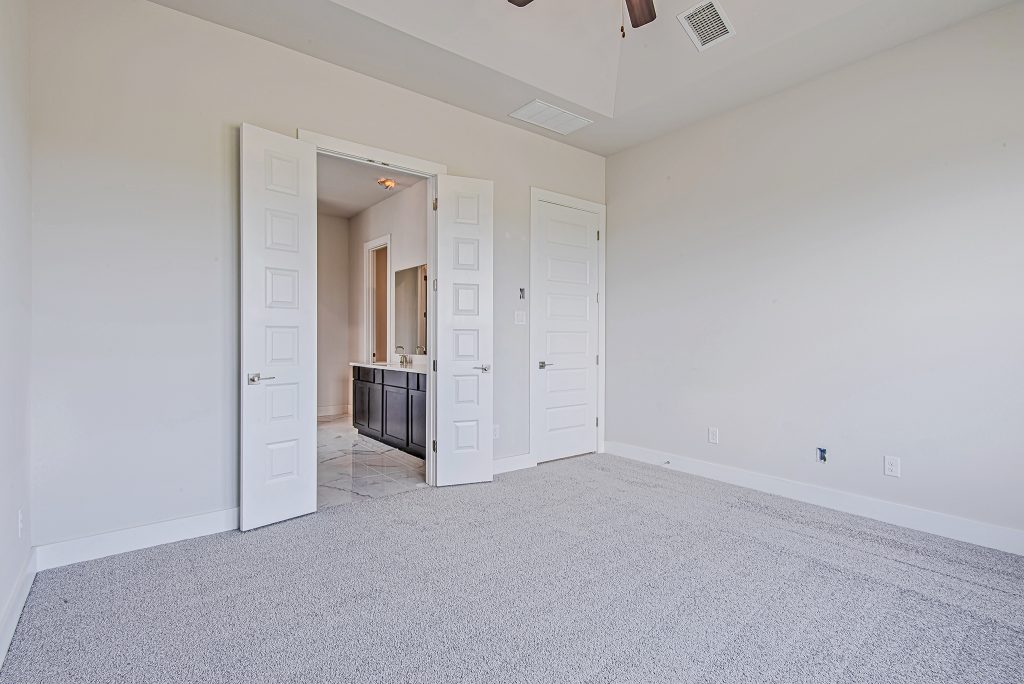 Empty bedroom with light gray carpet and white walls, featuring double doors leading to a bathroom with dark cabinets and a large mirror. The room has a high ceiling and a single closed door on the right wall.