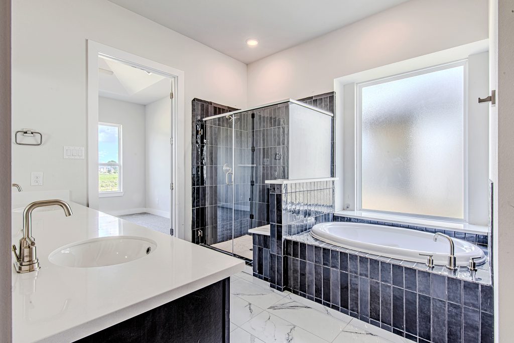 Modern bathroom with white walls, a large glass-enclosed shower, a built-in bathtub with dark tile surround, a white countertop with sink, and a frosted window letting in natural light.