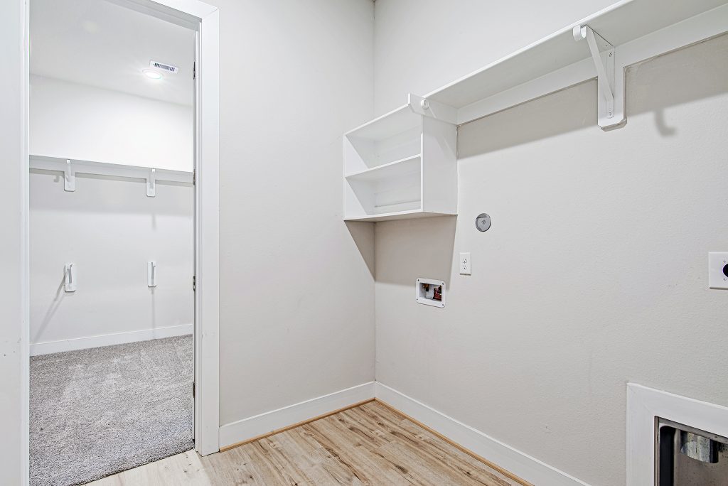A bright, empty laundry room with light wood flooring, white walls, built-in white shelves, and hook-ups for a washer and dryer. An open doorway leads to a carpeted closet with shelves and hanging rods.