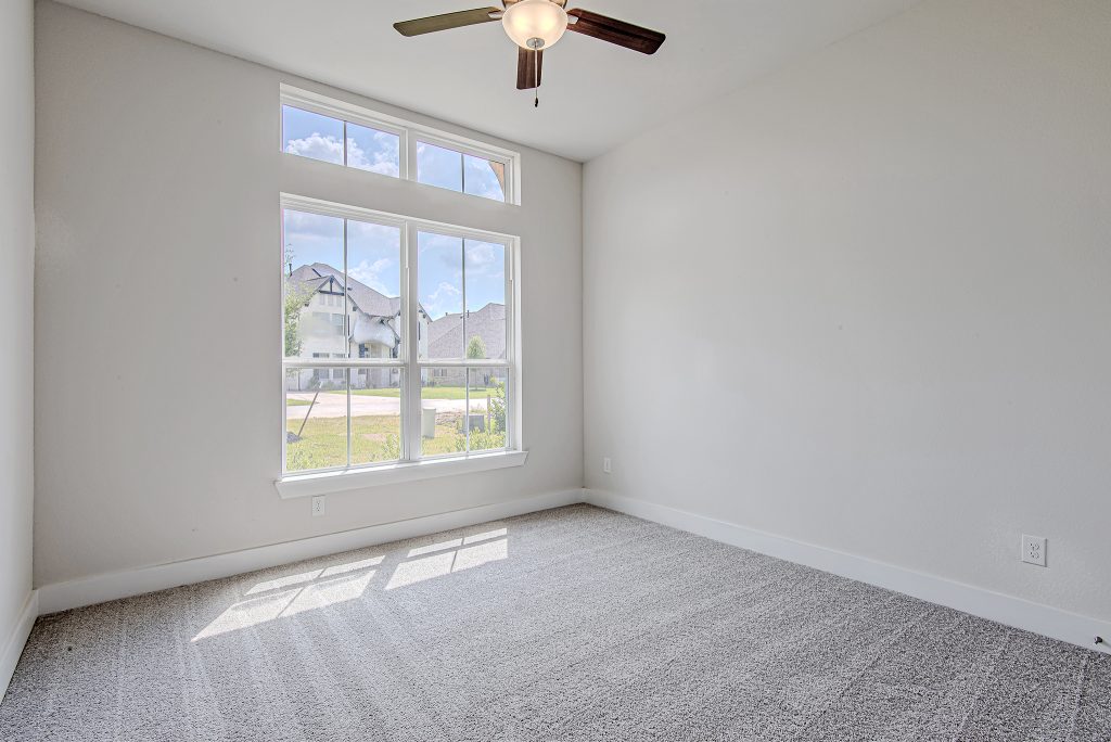 A bright, empty room with light gray carpet, white walls, a large window letting in natural light, and a ceiling fan with dark blades. Houses and a blue sky with clouds are visible through the window.