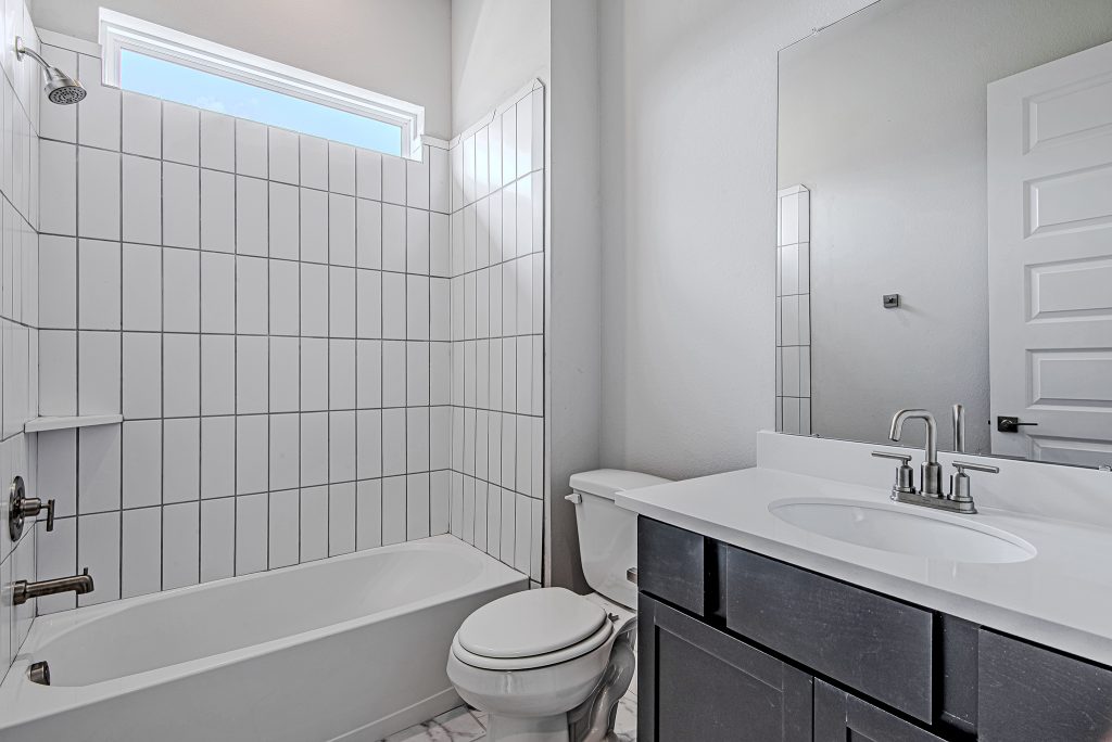 Modern bathroom with white tiled shower and bathtub, white toilet, dark vanity with white countertop, silver faucet, large mirror, and a window above the shower letting in natural light.