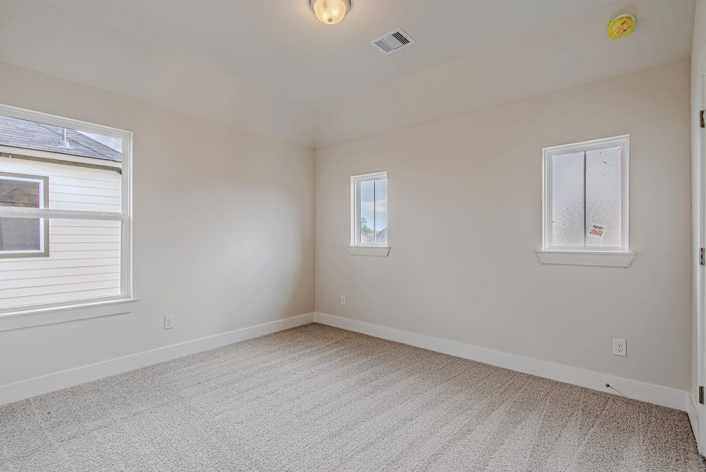 Empty, unfurnished room with beige carpet, white walls, a ceiling light, and three windows letting in natural light. The room has clean baseboards and an overall bright, neutral appearance.