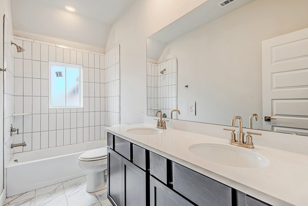 Modern bathroom with white tile walls, a bathtub with shower, a window, a double-sink vanity with dark cabinets, white countertops, and gold-colored faucets. White walls and tile floor complete the bright, clean space.