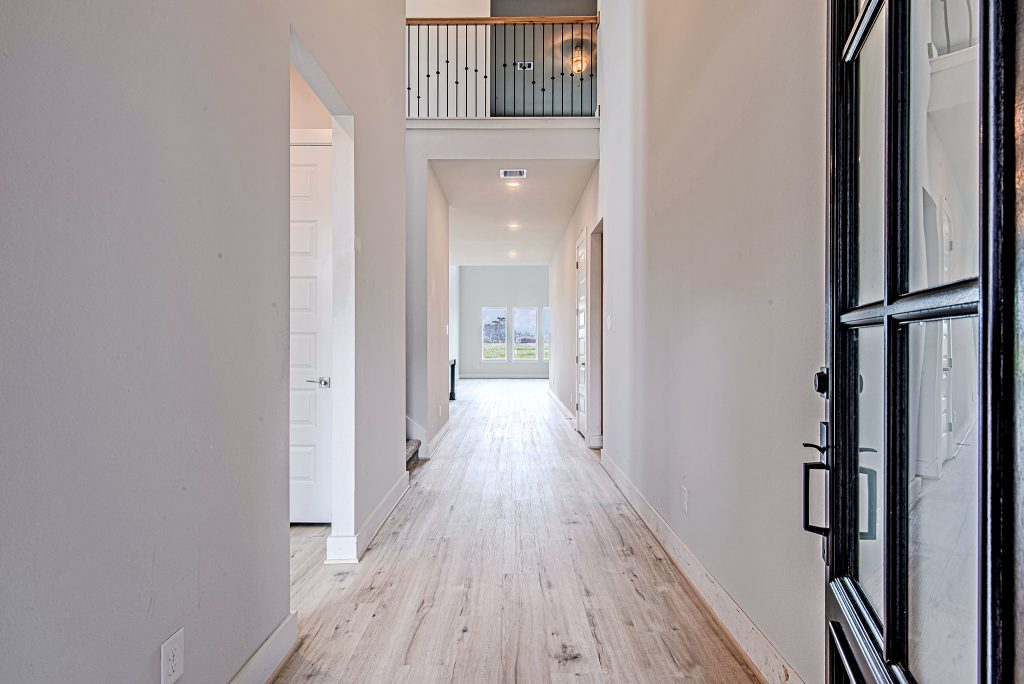 Bright, modern hallway with light wood flooring, white walls, and a black-framed glass door. The hallway leads to a living area with large windows and an upstairs railing is visible above.