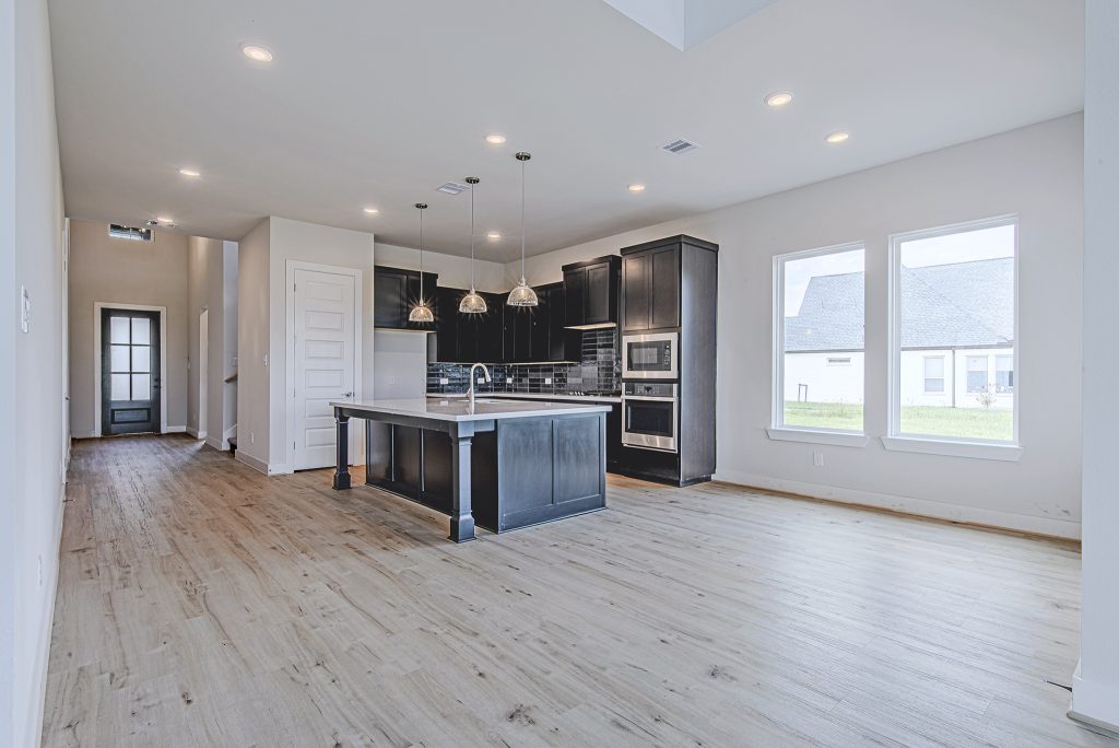 Modern kitchen with black cabinets, stainless steel appliances, an island with a sink, pendant lights, and light wood flooring. Large windows let in natural light, and the space opens to a hallway and adjacent rooms.