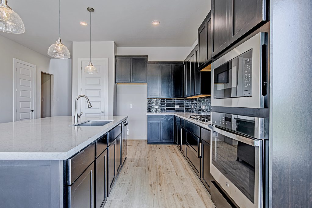 Modern kitchen with dark cabinets, stainless steel appliances, a built-in microwave and oven, white quartz countertops, glass pendant lights, and light wood flooring. The backsplash features a black and white tile pattern.