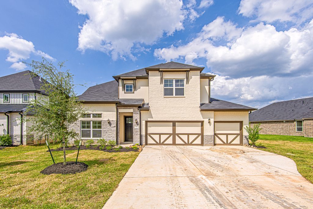 A modern two-story house with light brick exterior, dark roof, and a three-car garage. The driveway and small front yard with a young tree are visible under a partly cloudy blue sky.