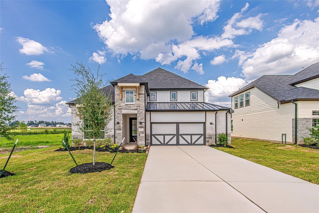 A modern two-story house with gray brick and stone exterior, black accents, a double garage, and a neatly landscaped front yard, set against a partly cloudy blue sky in a suburban neighborhood.