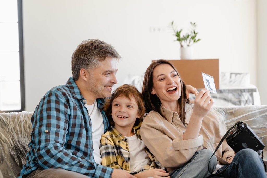 A smiling family of three—a man, a woman, and a young boy—sit closely together on a couch. The woman is holding a photograph and all three look happy and engaged.